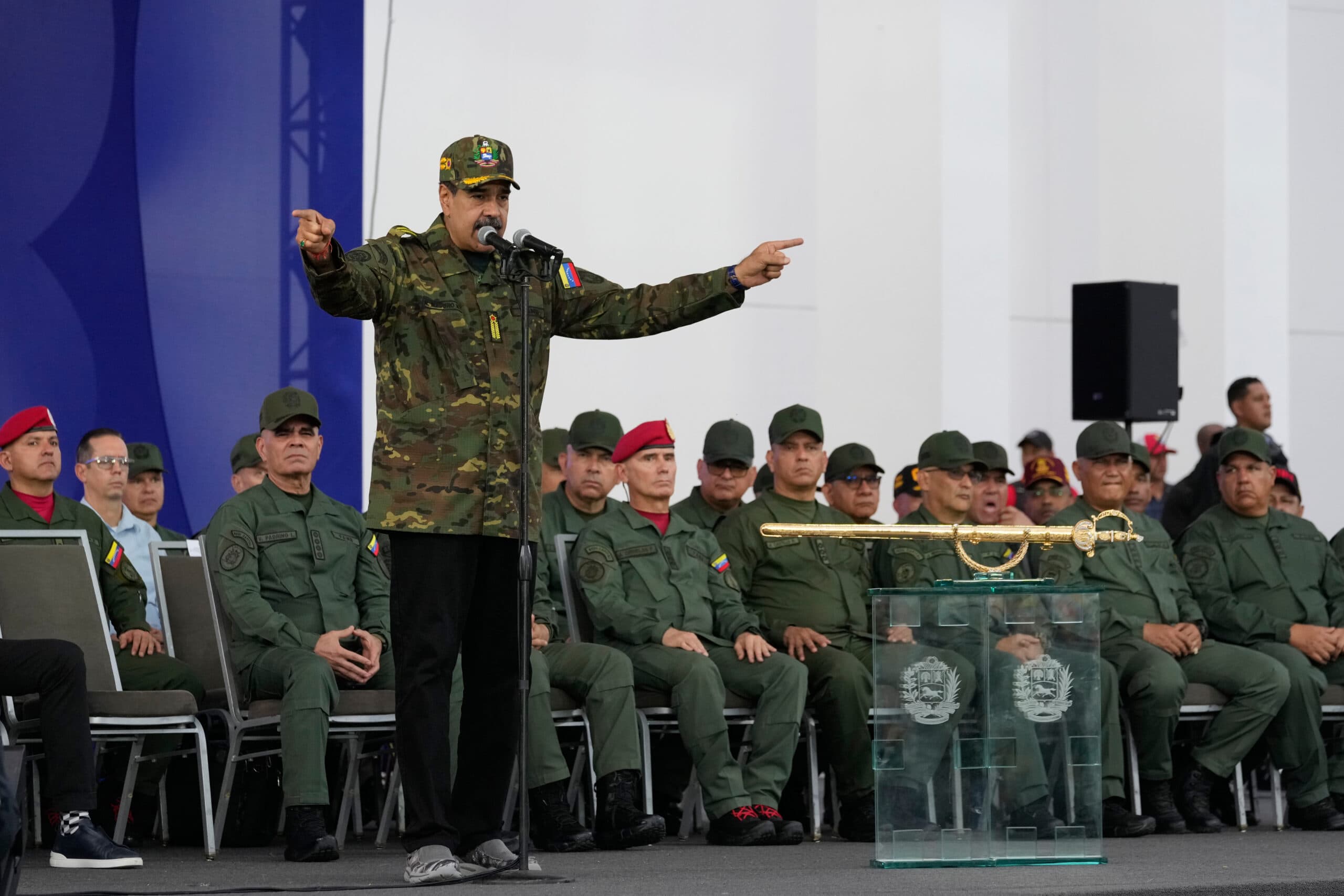 A man in a military uniform and cap speaks into a microphone while gesturing with both hands during a ceremony. He stands in front of a group of uniformed military personnel seated behind him. A ceremonial sword is displayed on a glass podium to his left.