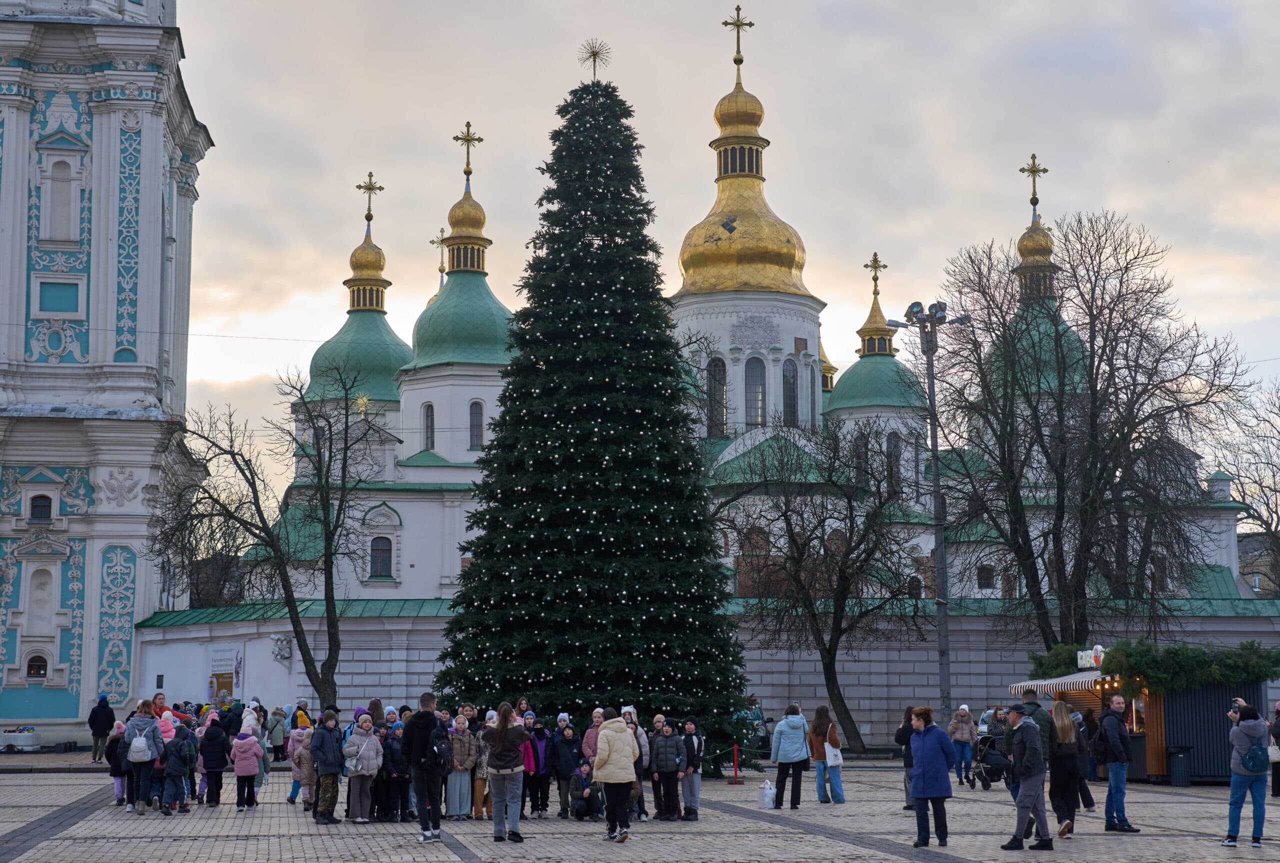 A group of people stands near a large decorated Christmas tree in front of a historic building with multiple green domes and golden crosses.