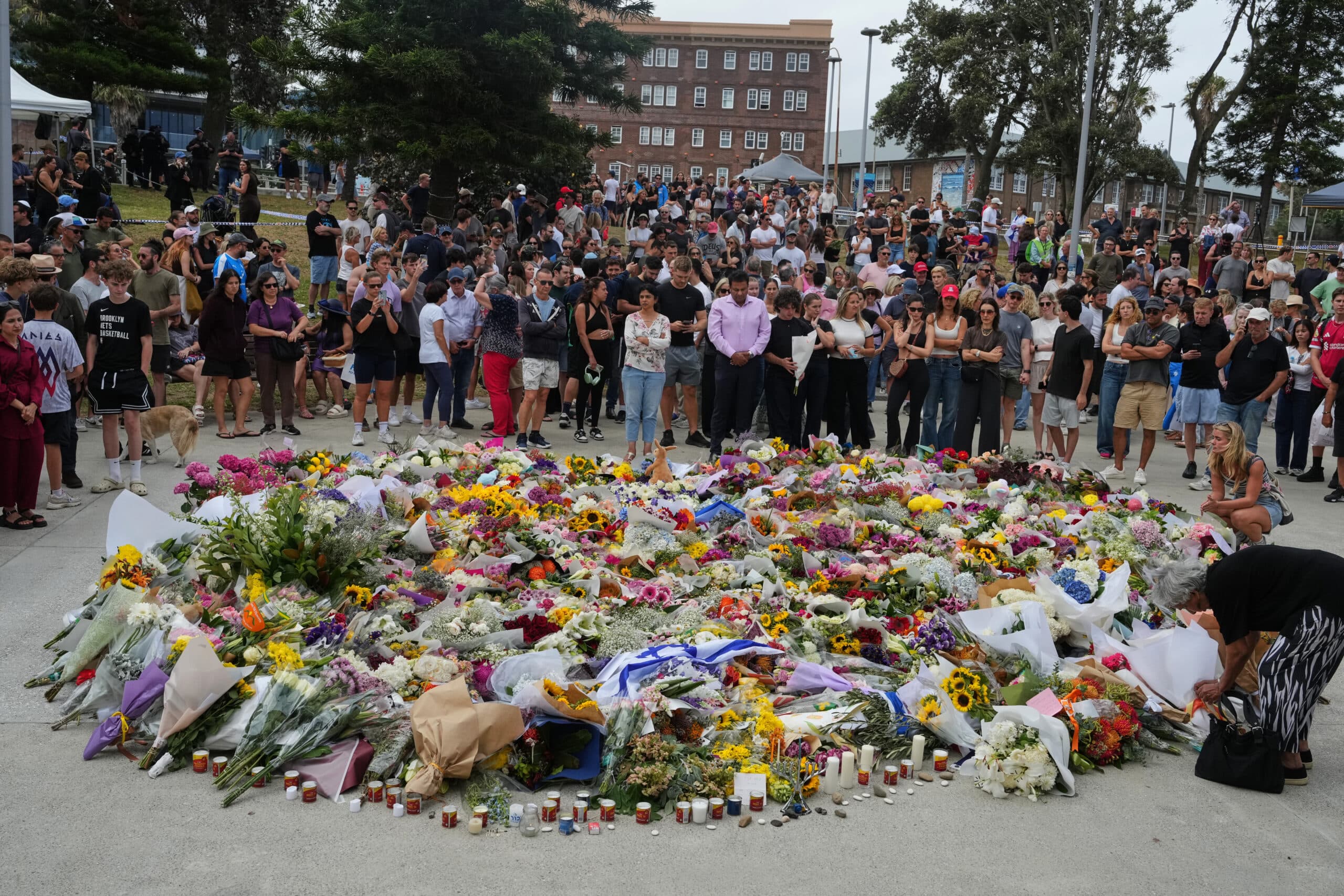 A large gathering of people surrounds a memorial site filled with numerous colorful flowers and candles, with a solemn atmosphere.