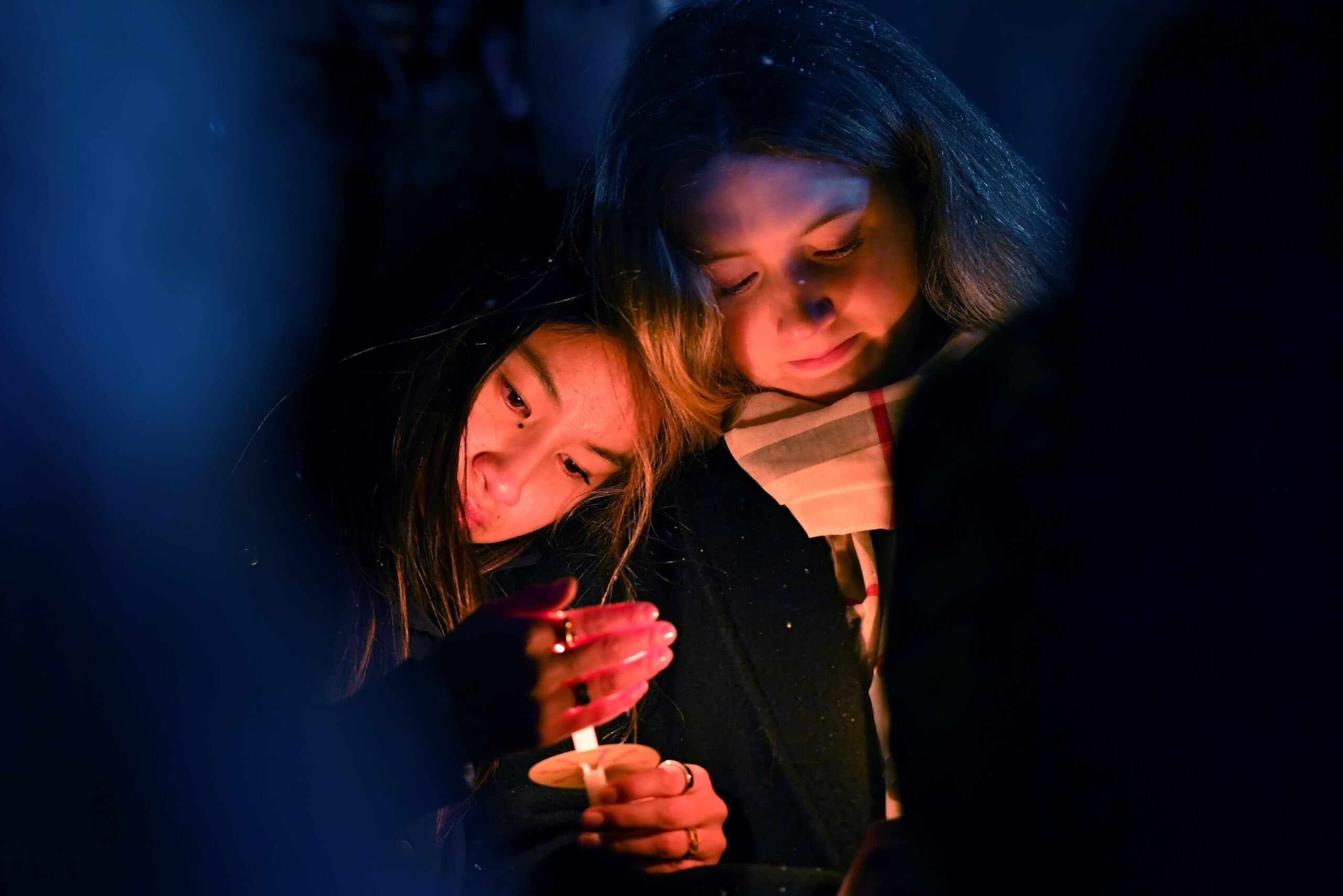 Two people are standing closely together, holding candles during a vigil or remembrance event, illuminated by the soft glow of candlelight in a dimly lit setting.