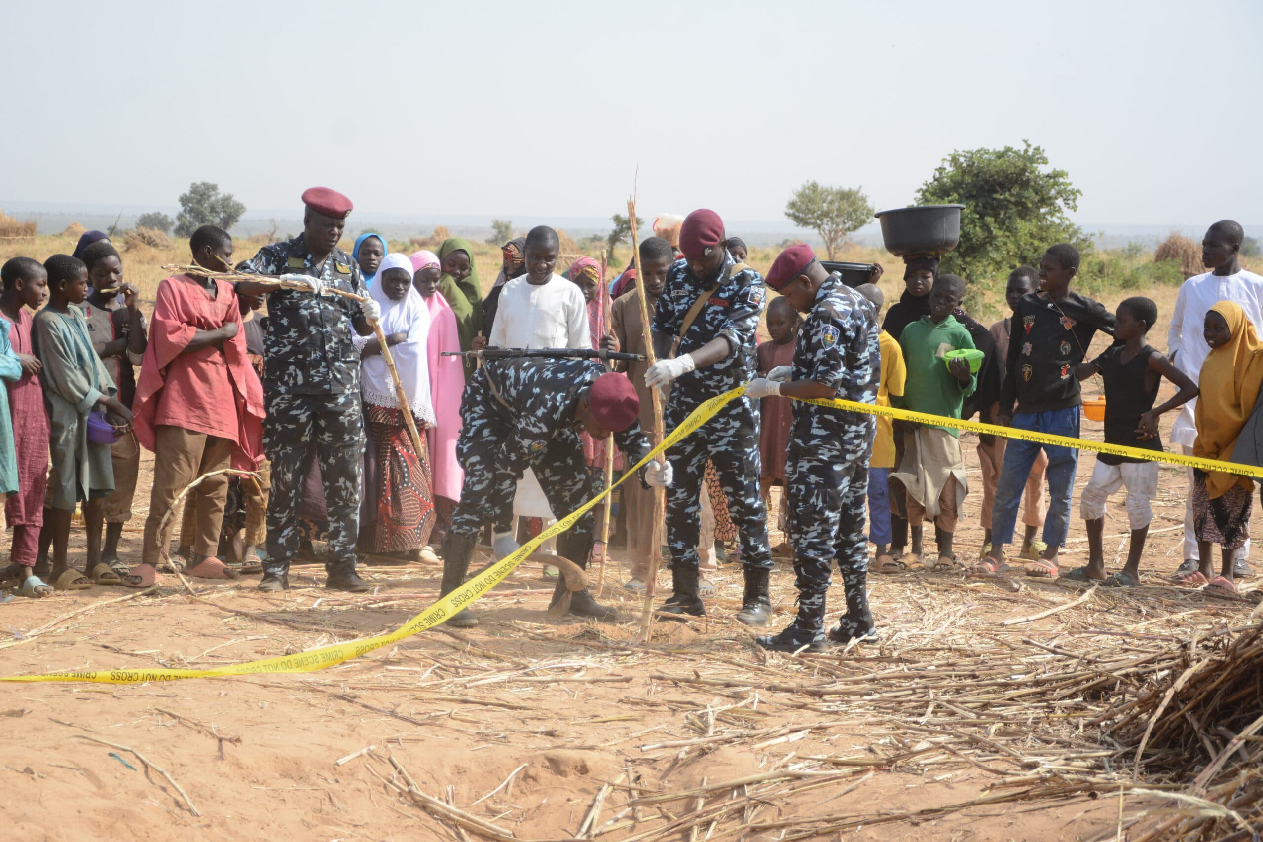A group of uniformed officers in camouflage, wearing maroon berets, work together to secure a crime scene with yellow tape, while a crowd of people, including adults and children, observe from behind the tape in a rural setting.