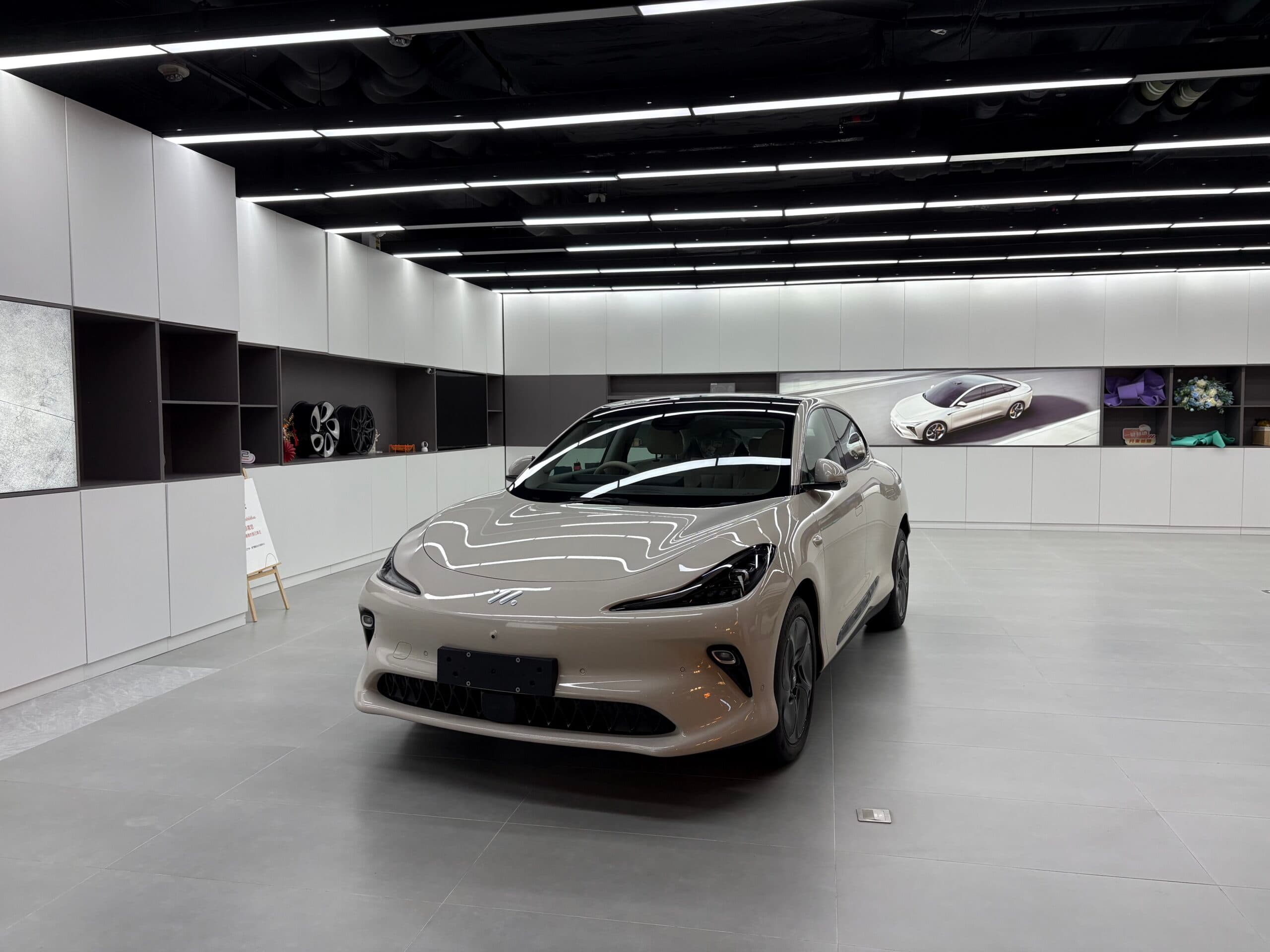 A sleek, modern car displayed in a showroom with a minimalist design, featuring white walls and shelving, and a large image of the car on display in the background.