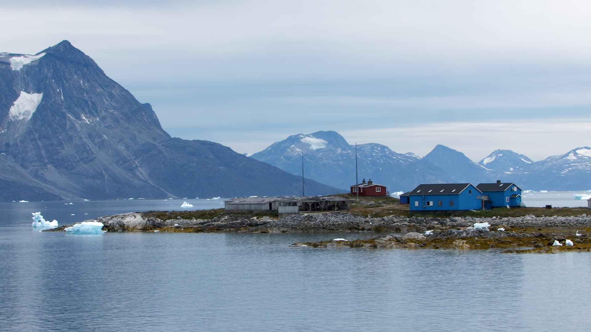 A small settlement with blue and red houses on a rocky islet surrounded by icy waters and distant mountains under a cloudy sky.