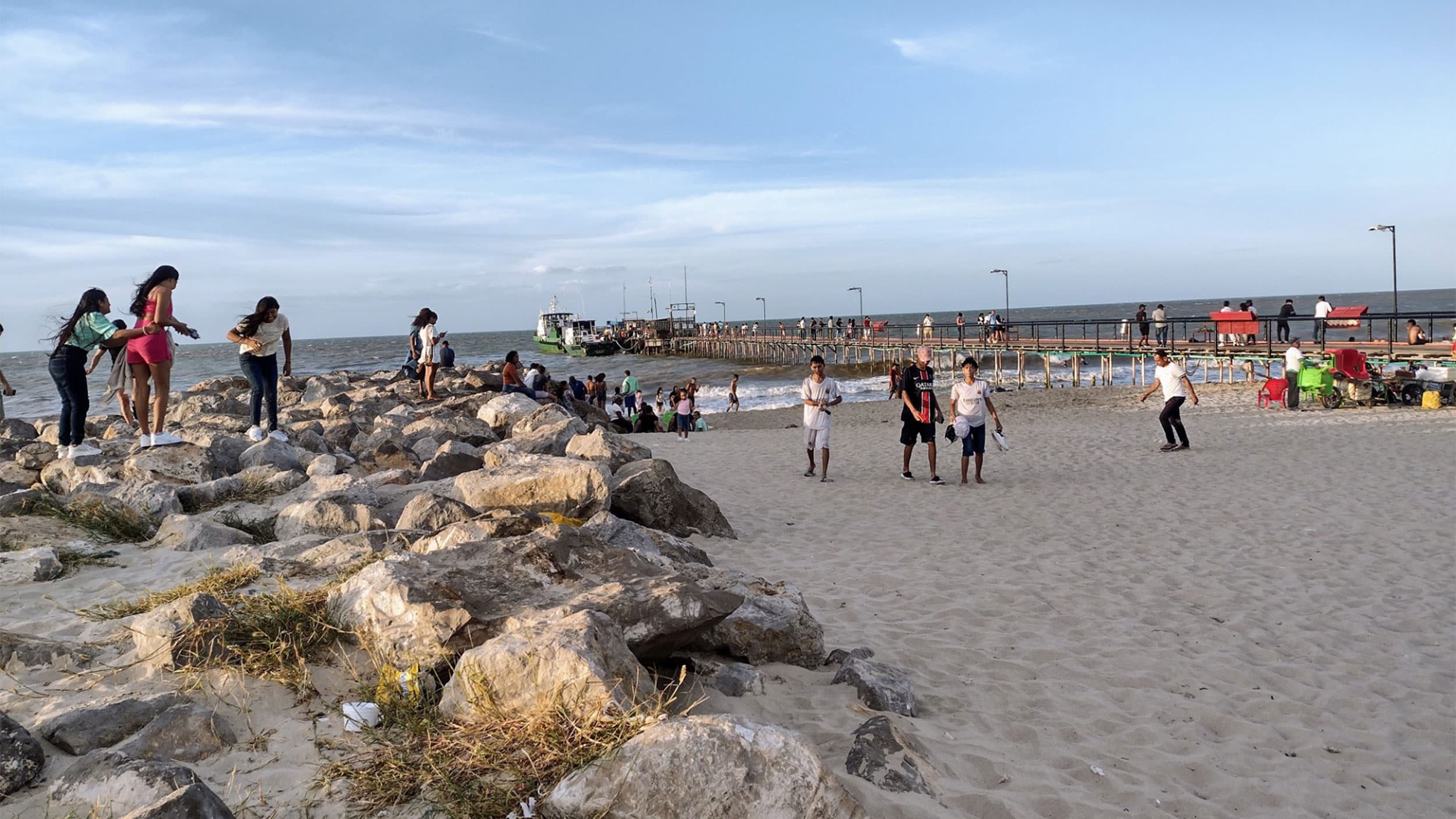 People walking and relaxing on a sandy beach with rocks and a pier extending into the ocean.