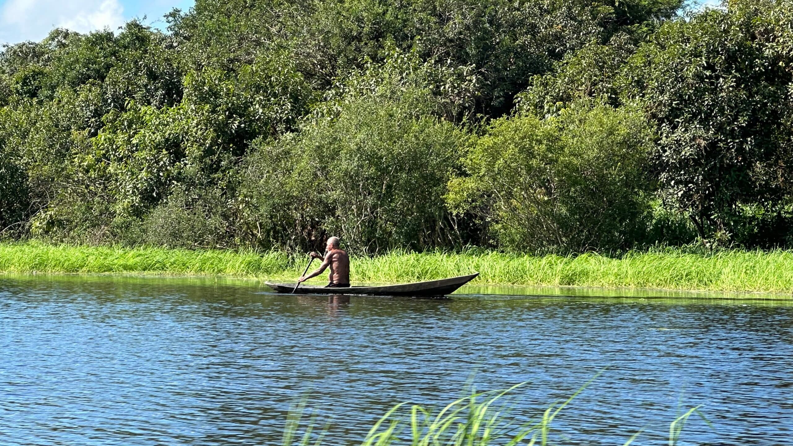 A person paddling a small wooden boat on a calm river surrounded by lush green vegetation.
