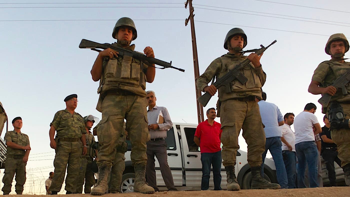 A group of soldiers in military uniforms and helmets stand guard with rifles in hand, with civilians and a vehicle visible in the background, under a clear sky.