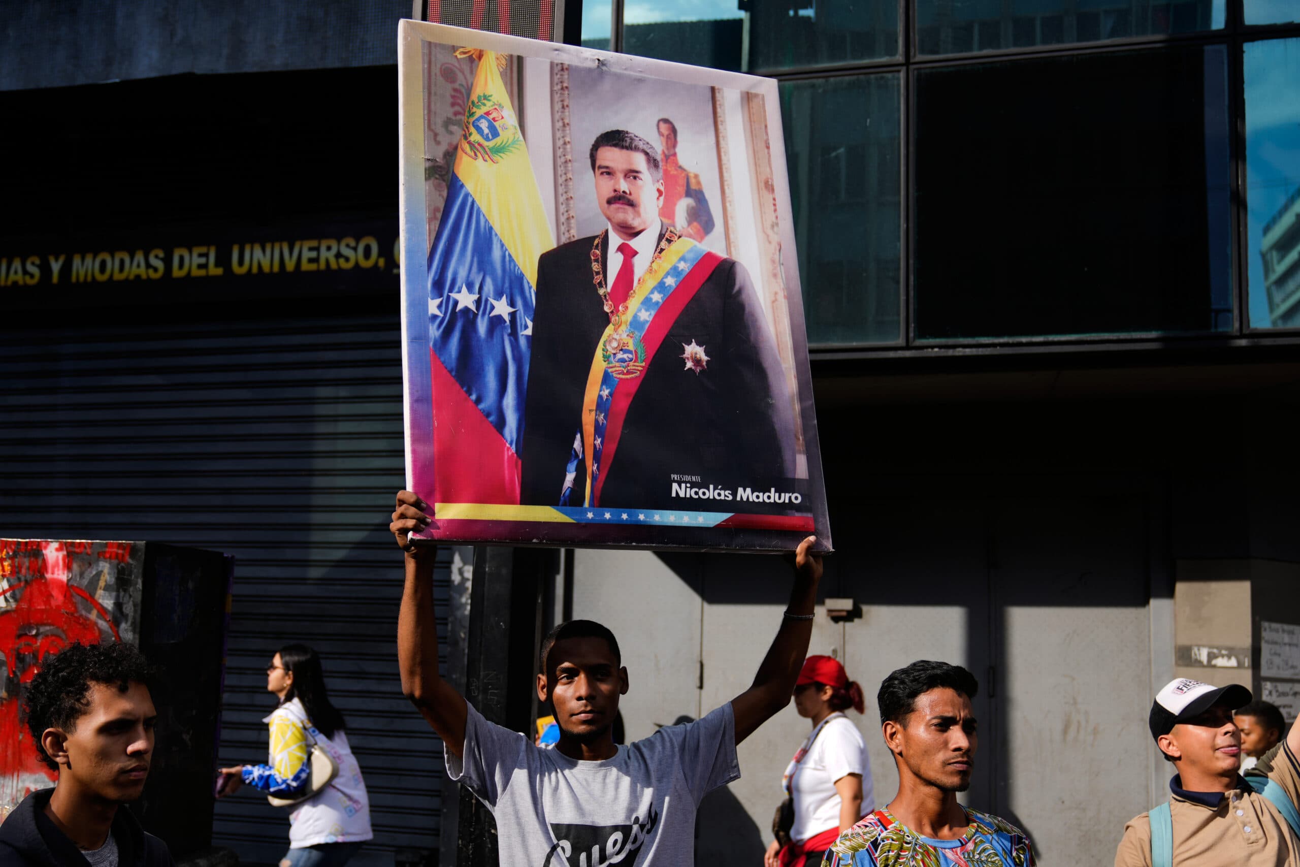 A man holds up a large poster of a person wearing a suit and sash, standing in front of a Venezuelan flag. Several people are visible around him on a street.