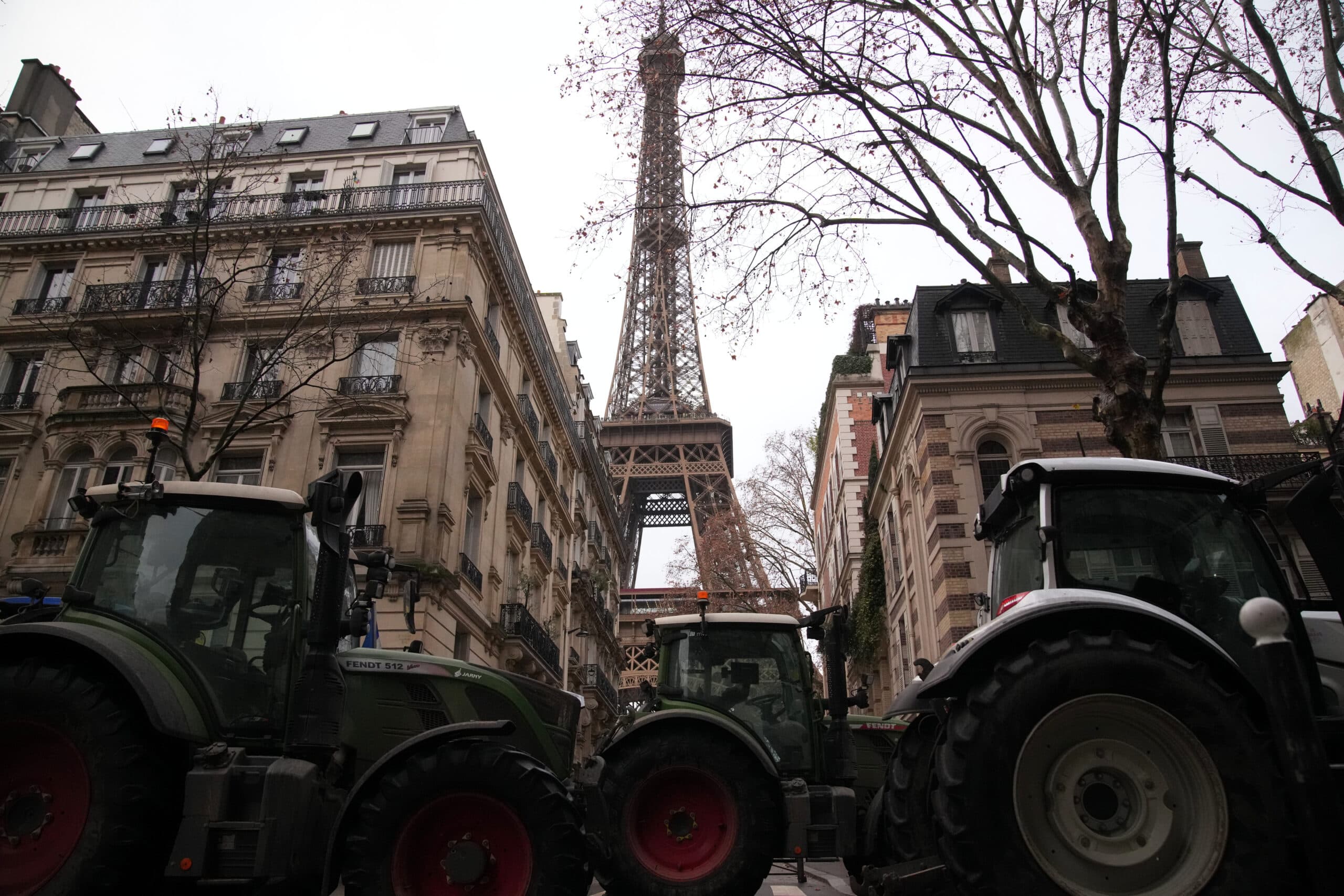 Tractors parked on a street in Paris with the Eiffel Tower visible in the background, framed by historic buildings and bare trees.