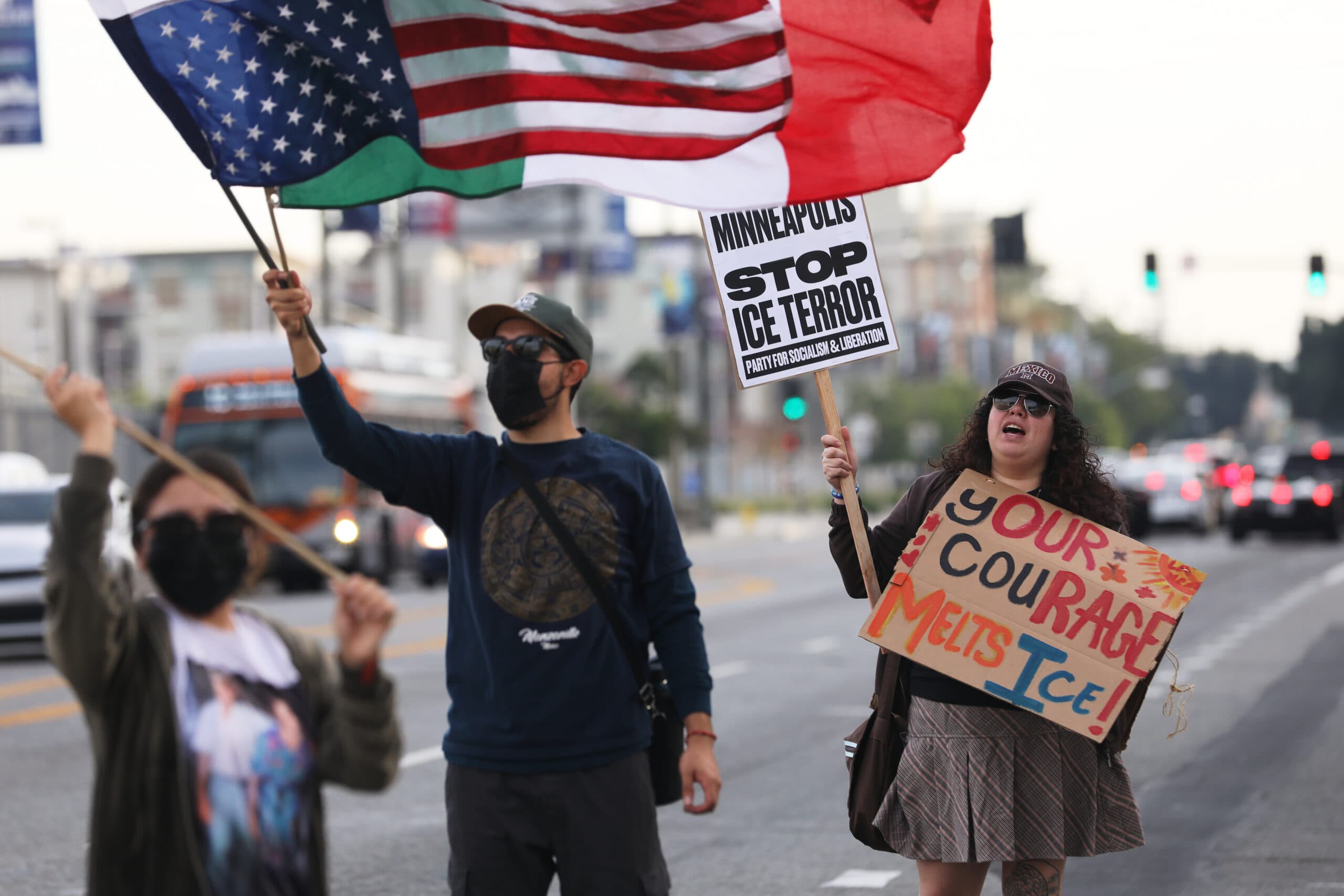 Three masked protesters stand on a street with blurred traffic in the background, holding signs and flags. One person holds an American flag and a sign that reads 'MINNEAPOLIS STOP ICE TERROR PARTY FOR SOCIALISM & LIBERATION.' Another holds a sign that says 'YOUR COURAGE MELTS ICE!'