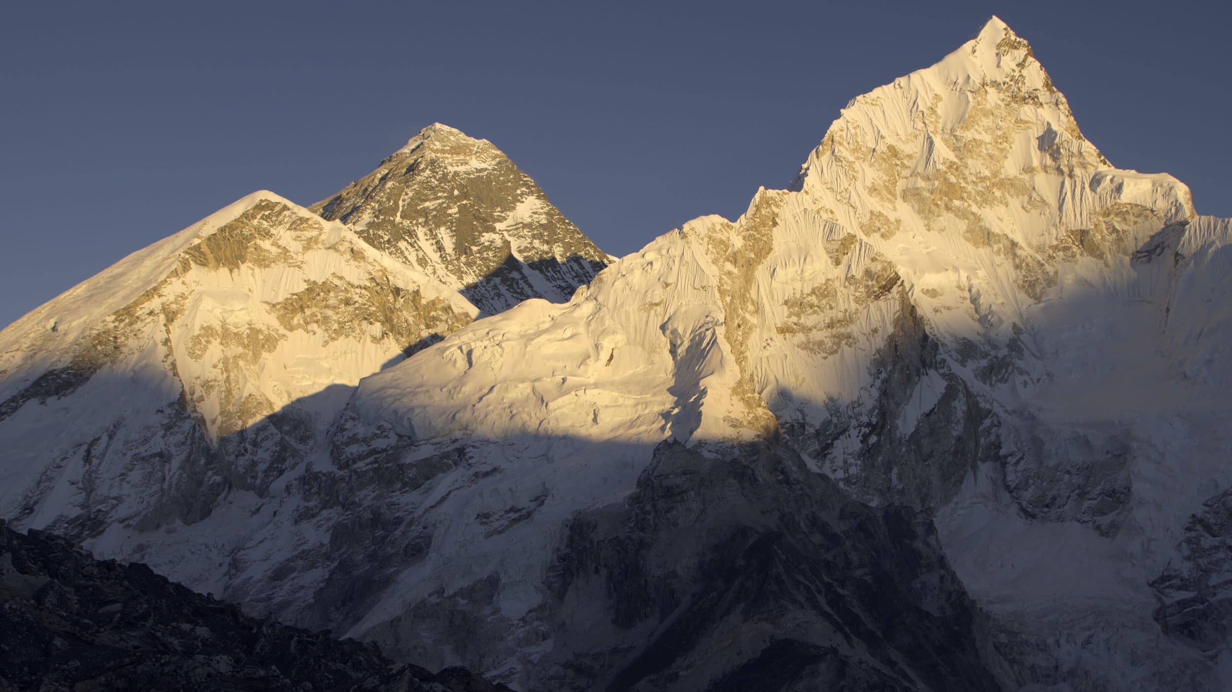 Snow-covered mountain peaks, including Mount Everest, illuminated by golden sunlight, casting shadows on the slopes under a clear sky.