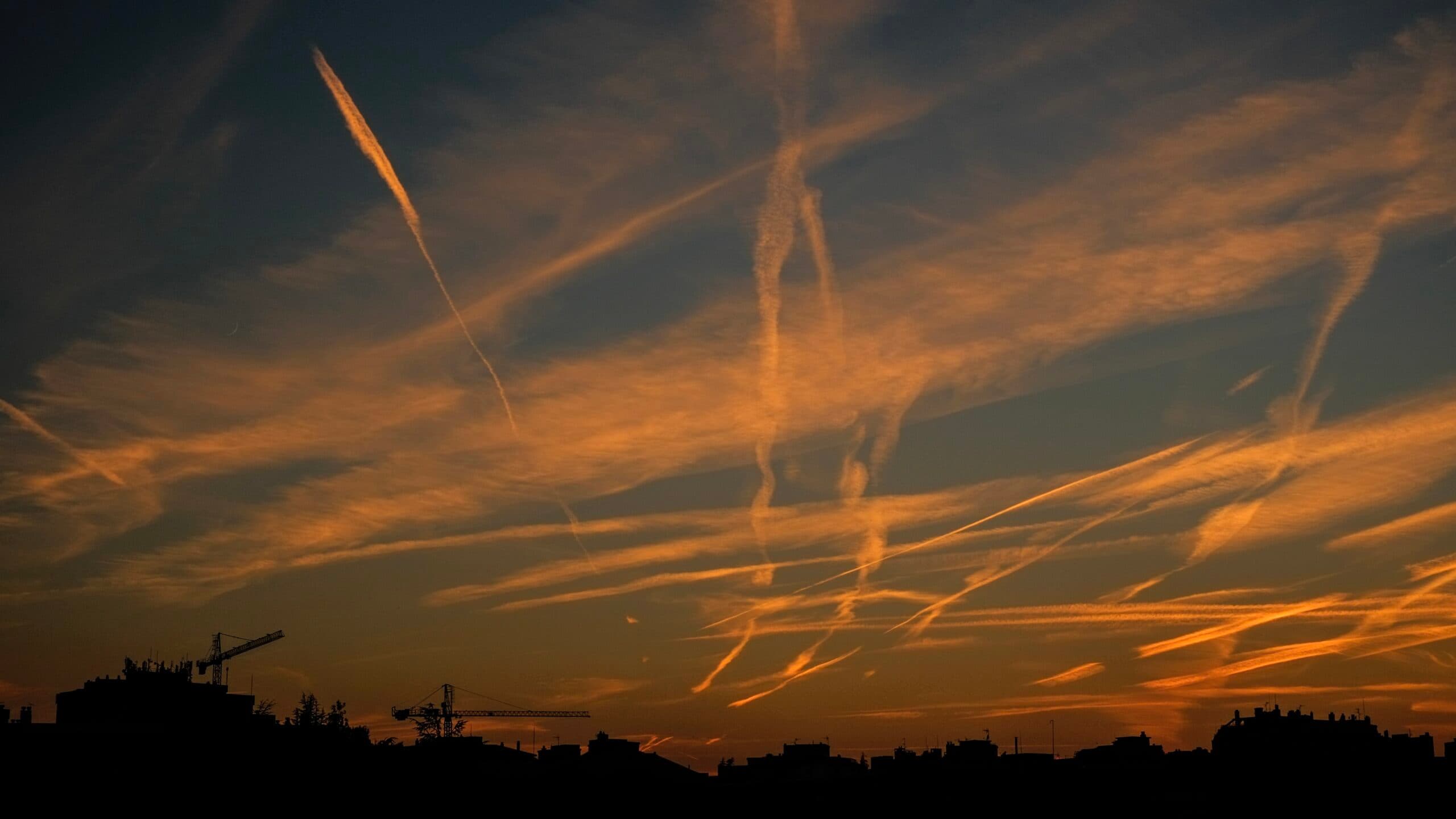 Silhouette of a city skyline with cranes against a sunset sky filled with orange contrails and wispy clouds.