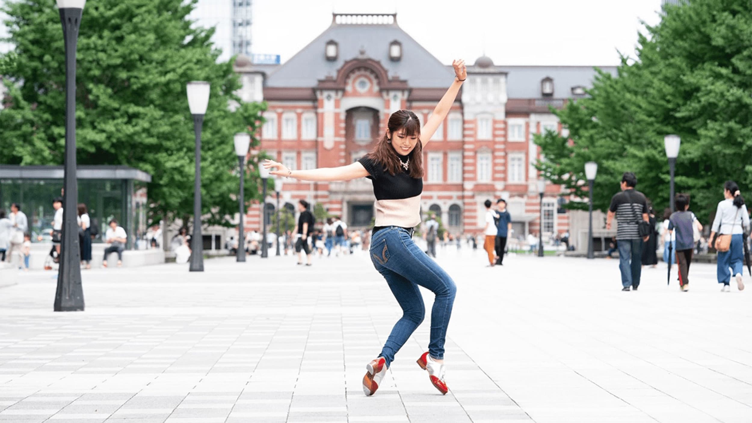 A woman in a black top and jeans performs a dynamic dance move on a plaza with a historic red brick building in the background. Other people are walking by and trees line the square.