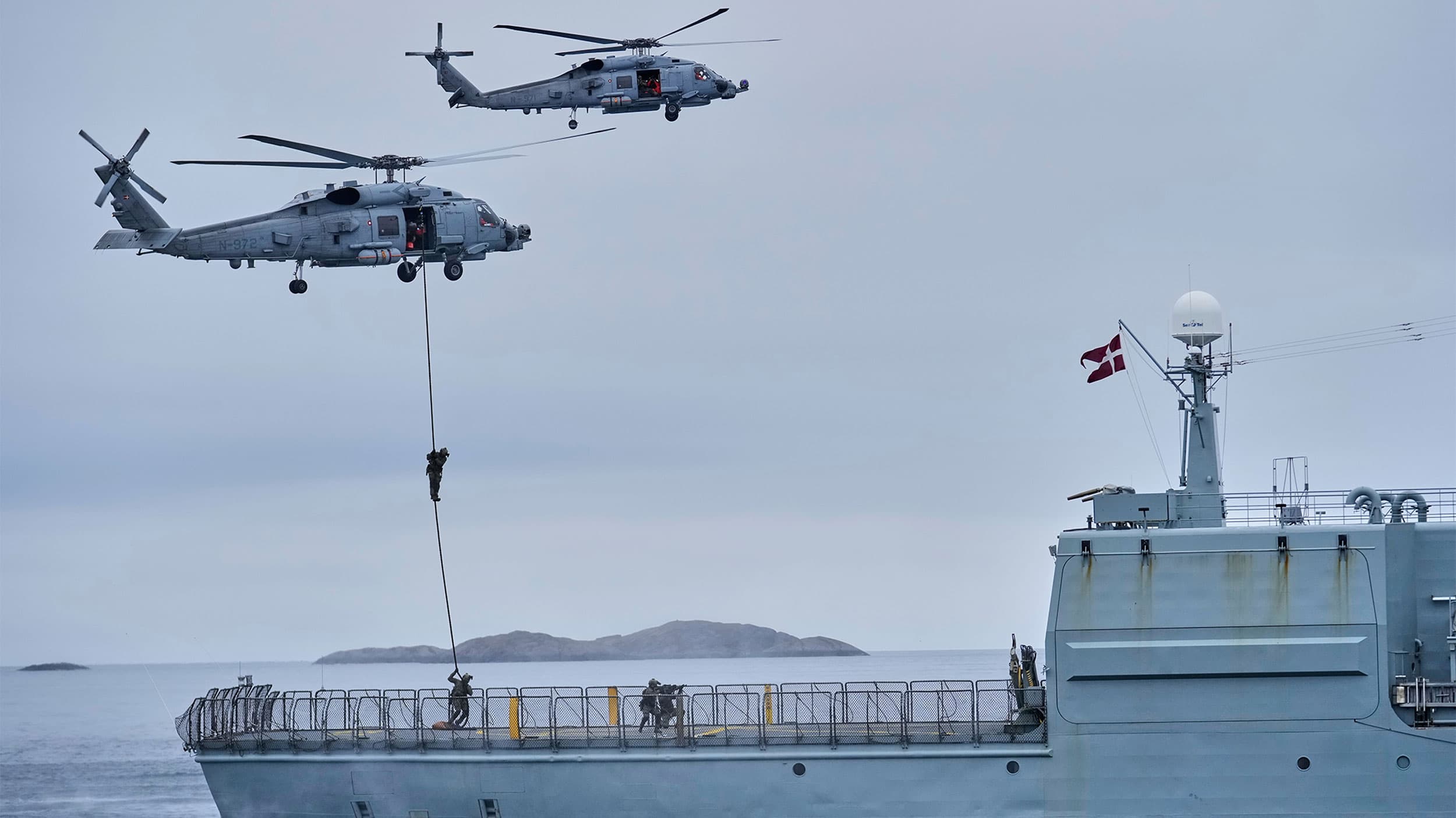 Two military helicopters hover above a ship, with one soldier rappelling down from a helicopter rope onto the deck, displaying a Danish flag.