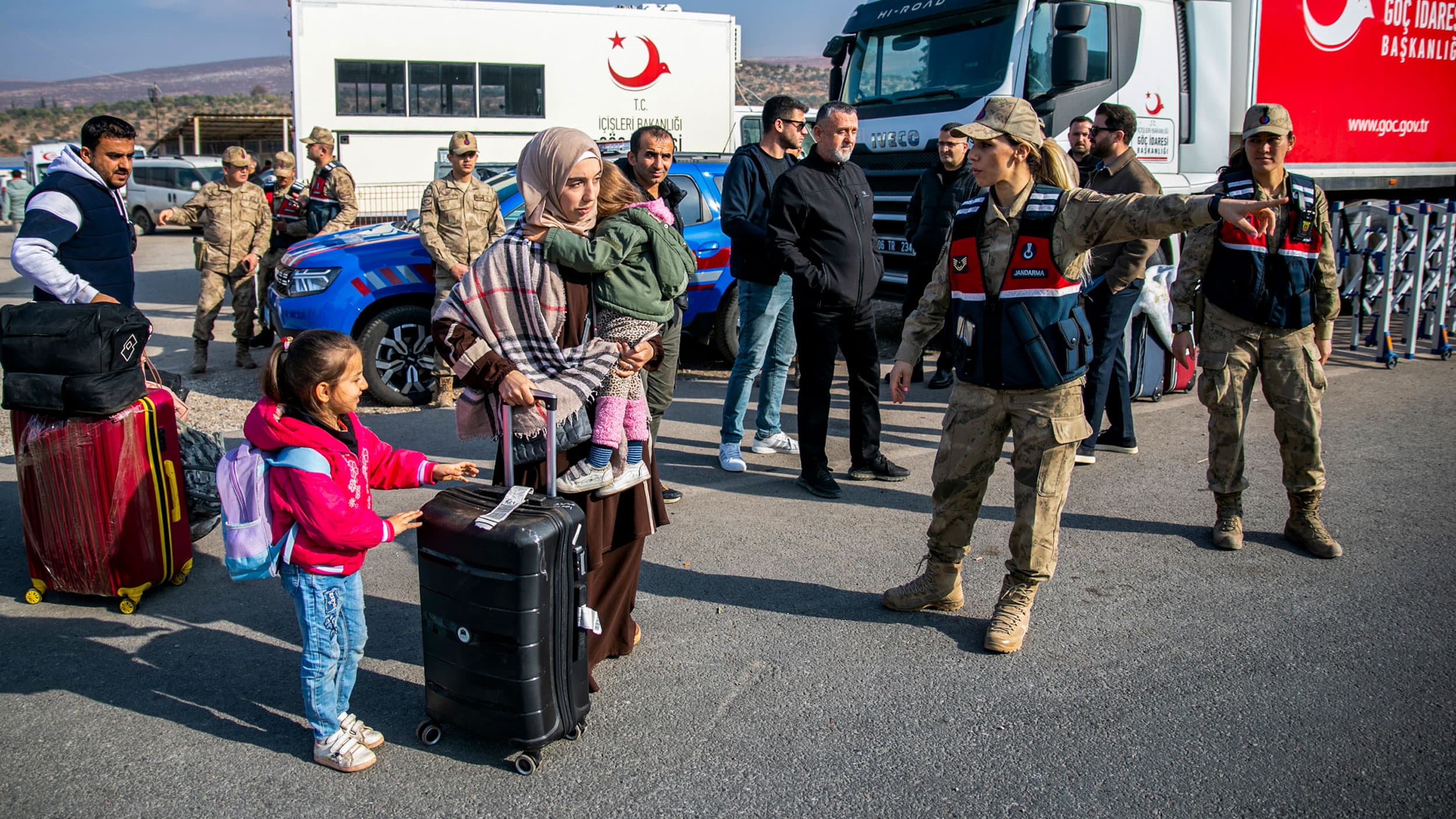 A group of people, including a woman holding a child and a young girl with a backpack, are seen at an outdoor location with military personnel and vehicles. The woman is wearing a headscarf and carrying luggage, and a soldier is directing the group. Military trucks and personnel are visible in the background.