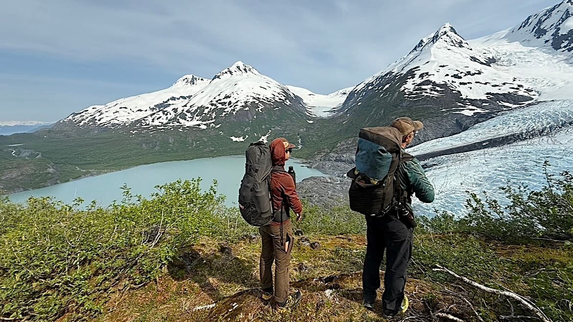 Two hikers with backpacks stand on a hill overlooking a glacier, snow-capped mountains, and a turquoise lake below, surrounded by lush green vegetation.