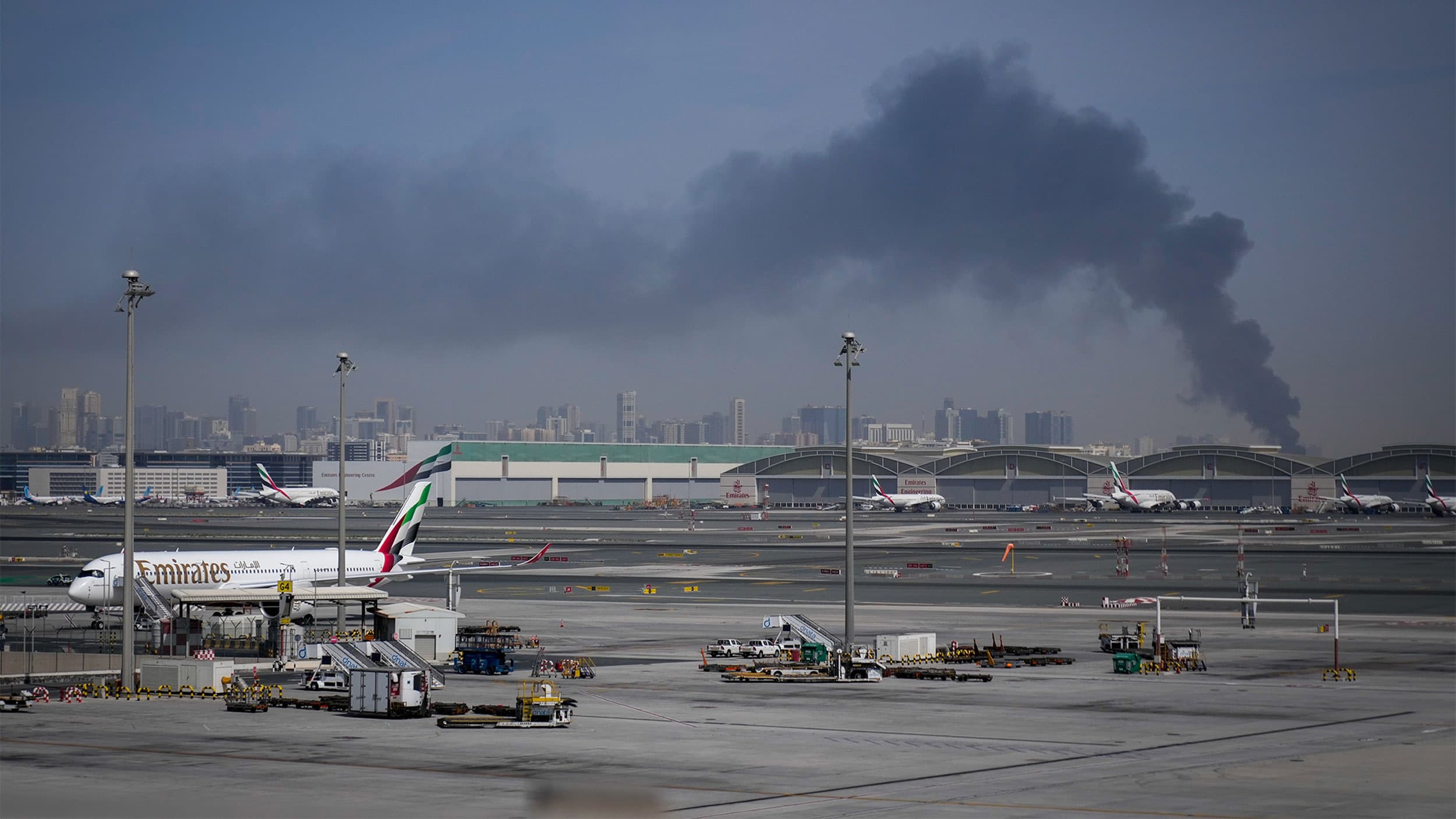 View of an airport with multiple airplanes parked on the tarmac, set against a city skyline in the background and a large plume of dark smoke rising into the sky.