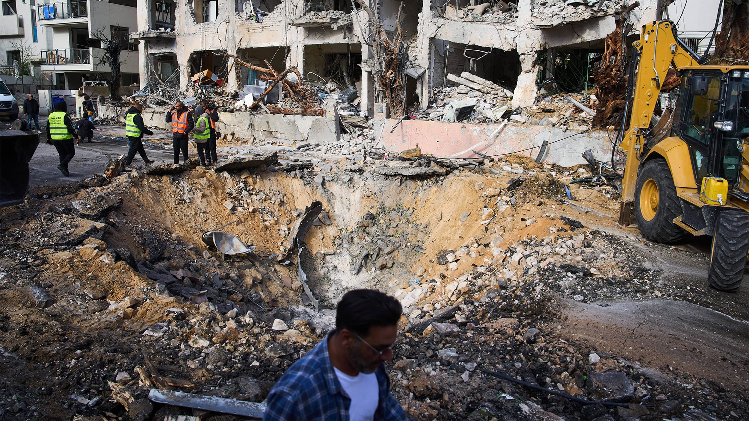 Rescue workers in high-visibility vests stand near a large crater caused by a missile strike in front of a heavily damaged building, with construction equipment nearby.