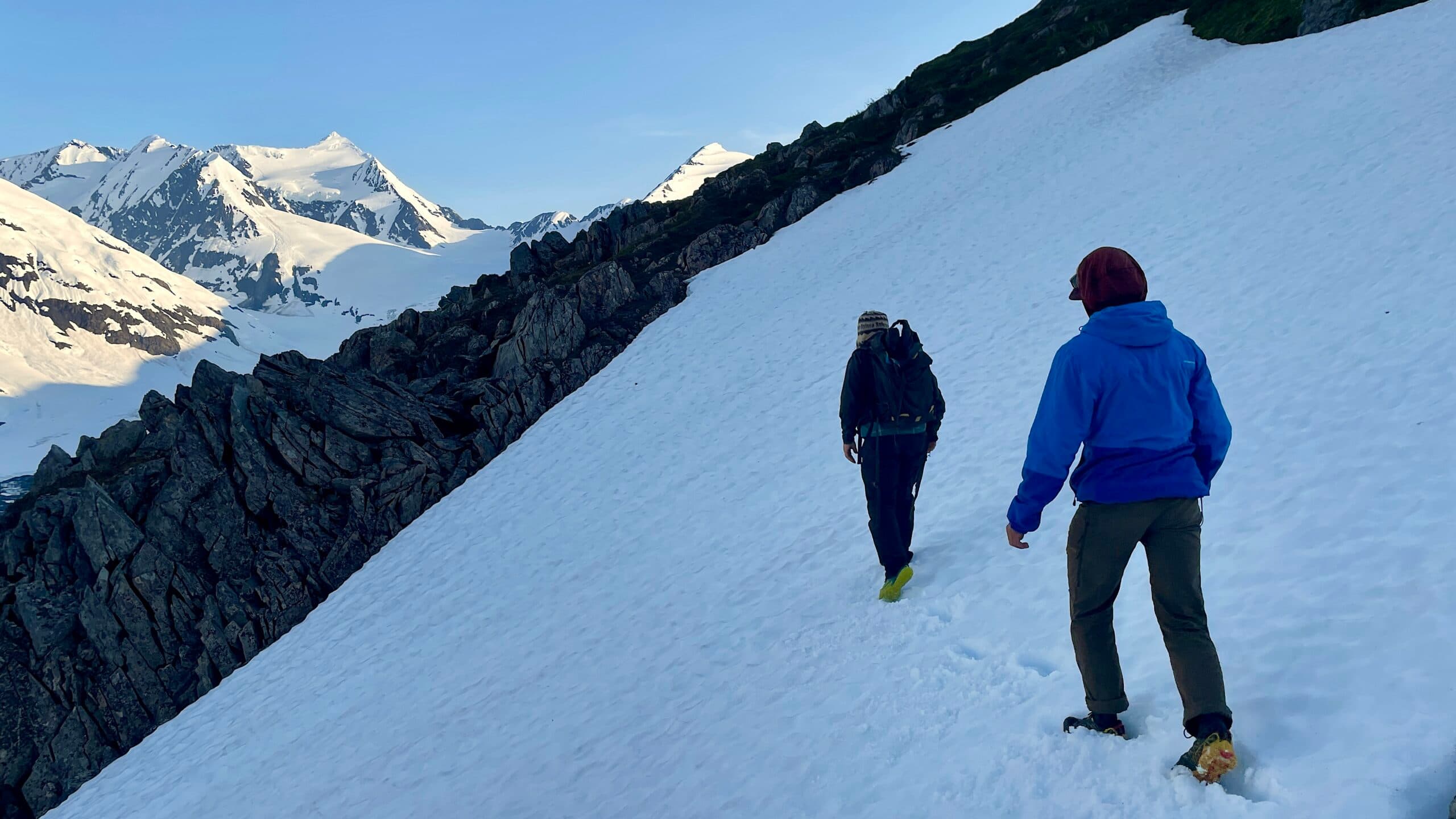 Two hikers wearing cold-weather gear traverse a snowy mountain slope, with snow-covered peaks visible in the background under a clear blue sky.
