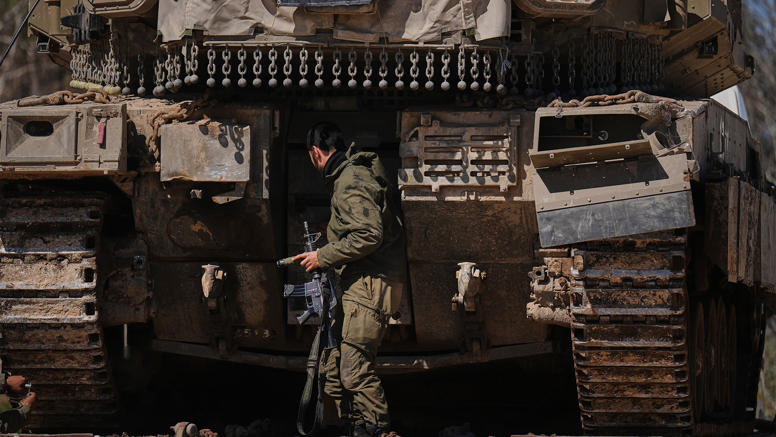 A soldier in green camouflage uniform stands beside a large military tank, holding a rifle and examining its exterior in a dusty environment.