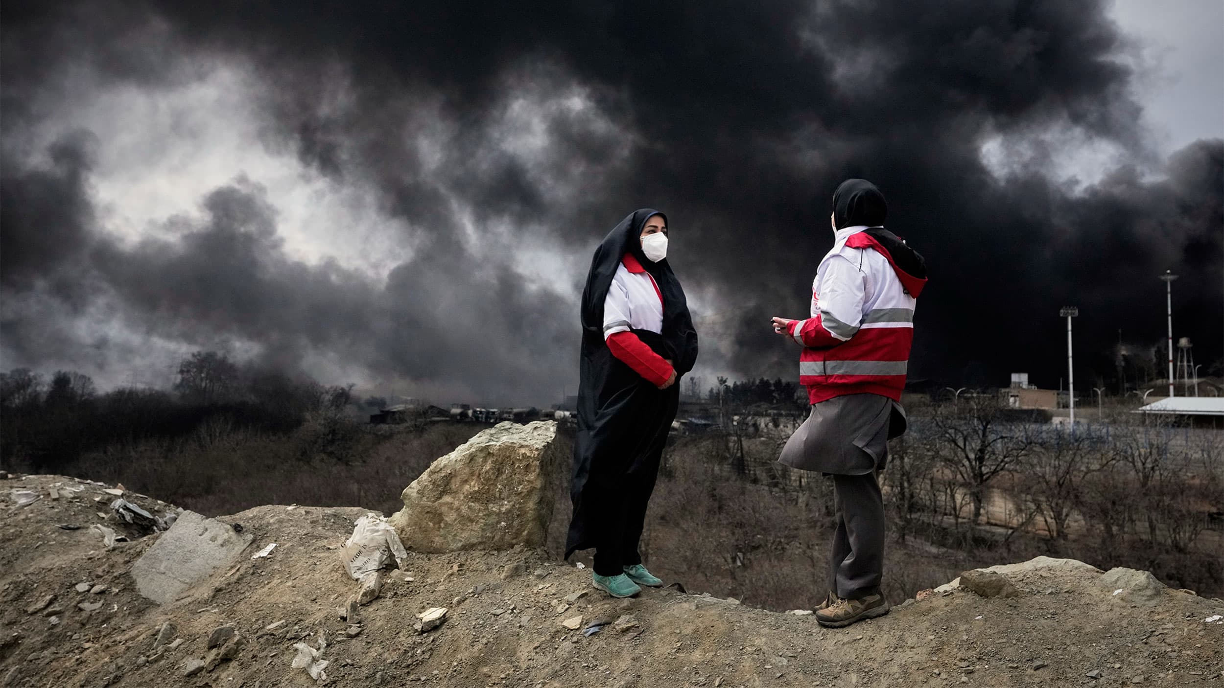 Two people wearing protective masks and Red Crescent uniforms stand on rocky terrain with a backdrop of thick black smoke and clouds.