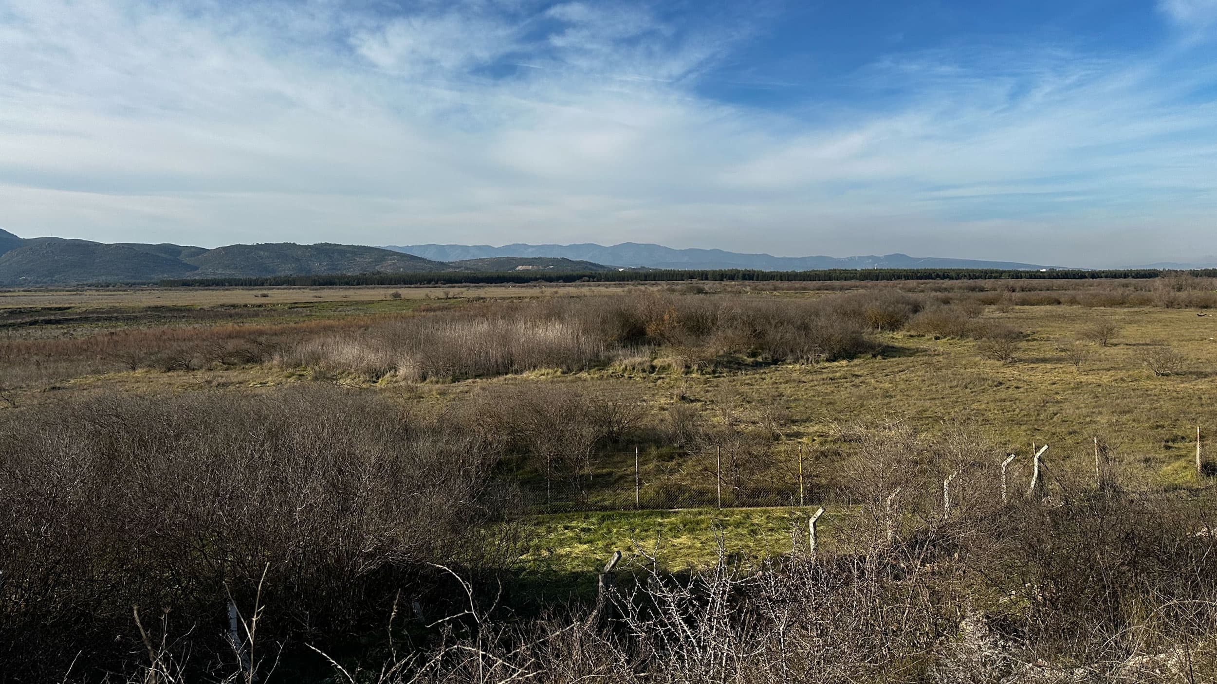 A vast rural landscape featuring dry grasslands with scattered shrubs and bushes, set against a backdrop of distant hills and a clear blue sky.