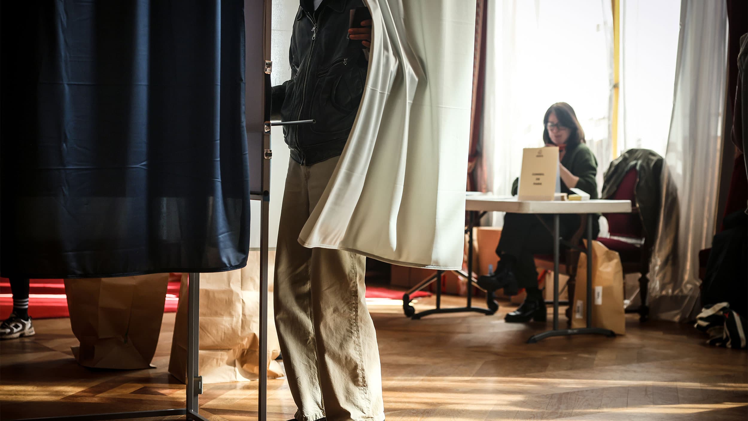 A person standing partially behind a voting booth curtain with another person sitting at a table nearby, likely at a polling station.