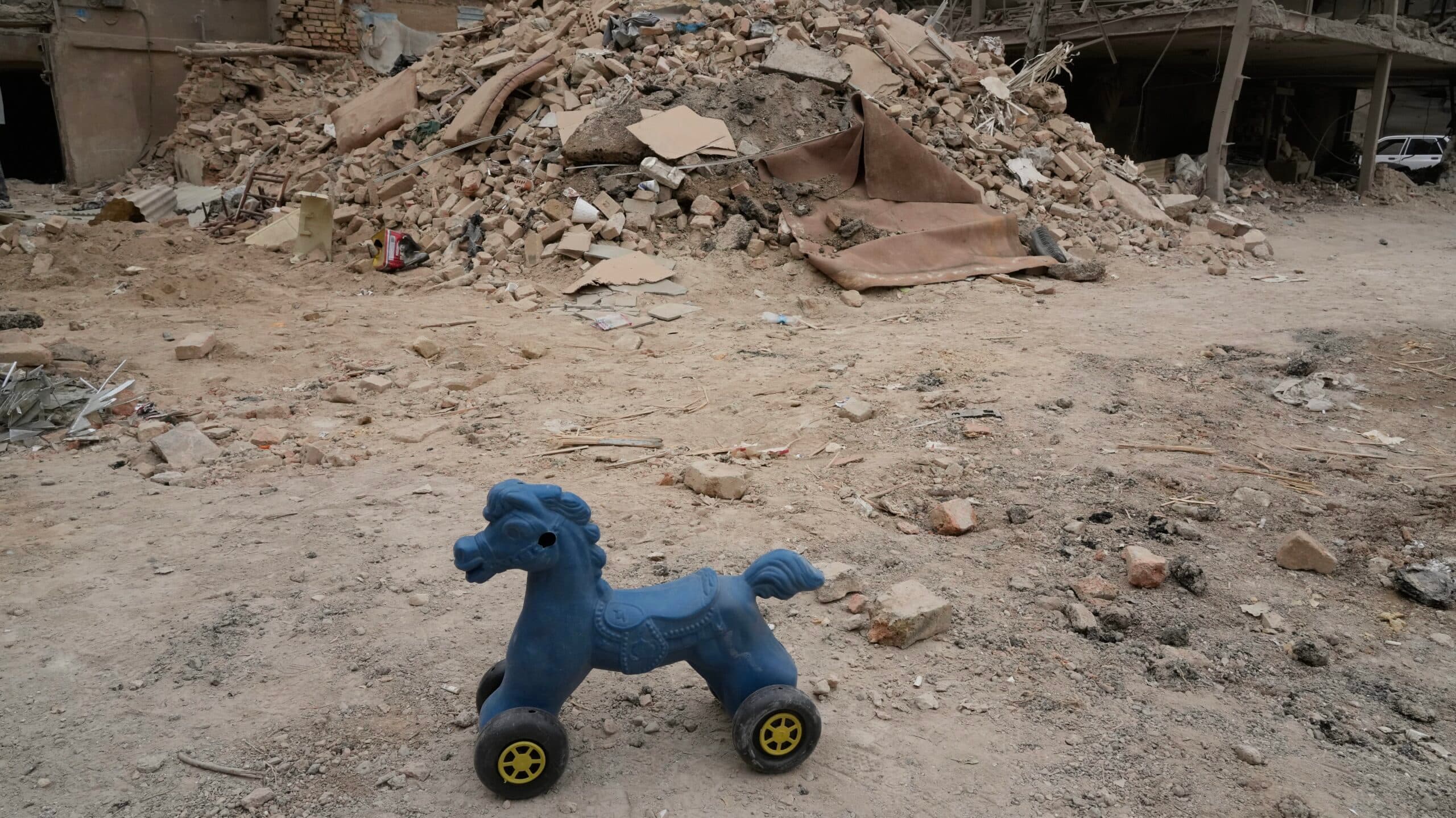 A blue plastic toy horse with wheels stands on a dirt ground in front of a pile of rubble and debris, including bricks and pieces of wood.