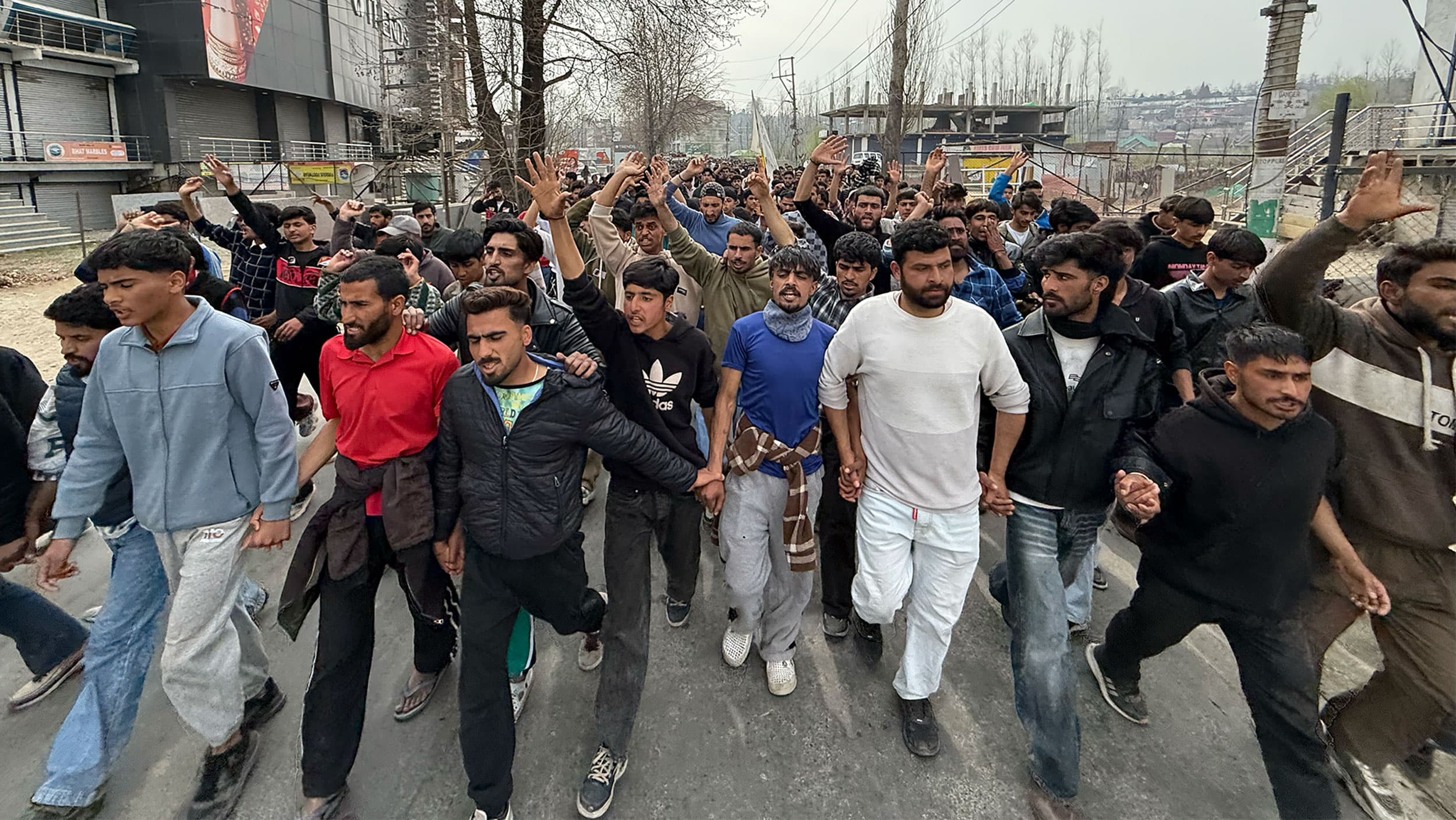 A large group of people walking in a demonstration, many with raised arms and hands clasped together, on a street lined with trees and buildings.