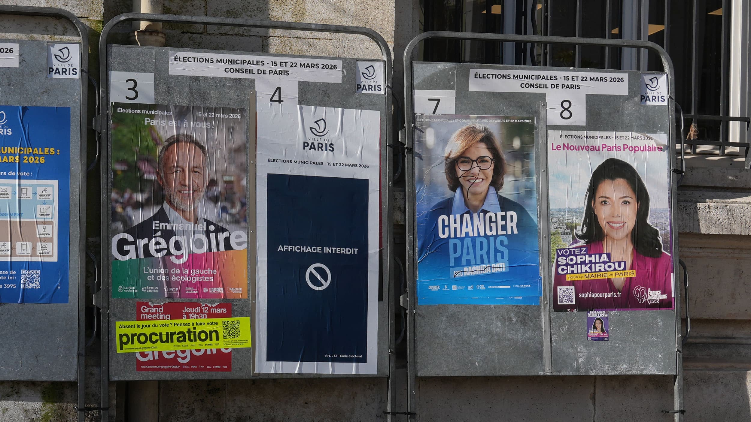 Election campaign posters displayed on metal boards in Paris, with signs for the 2026 municipal elections. Features images and names of three candidates, and a notice indicating prohibited postings.
