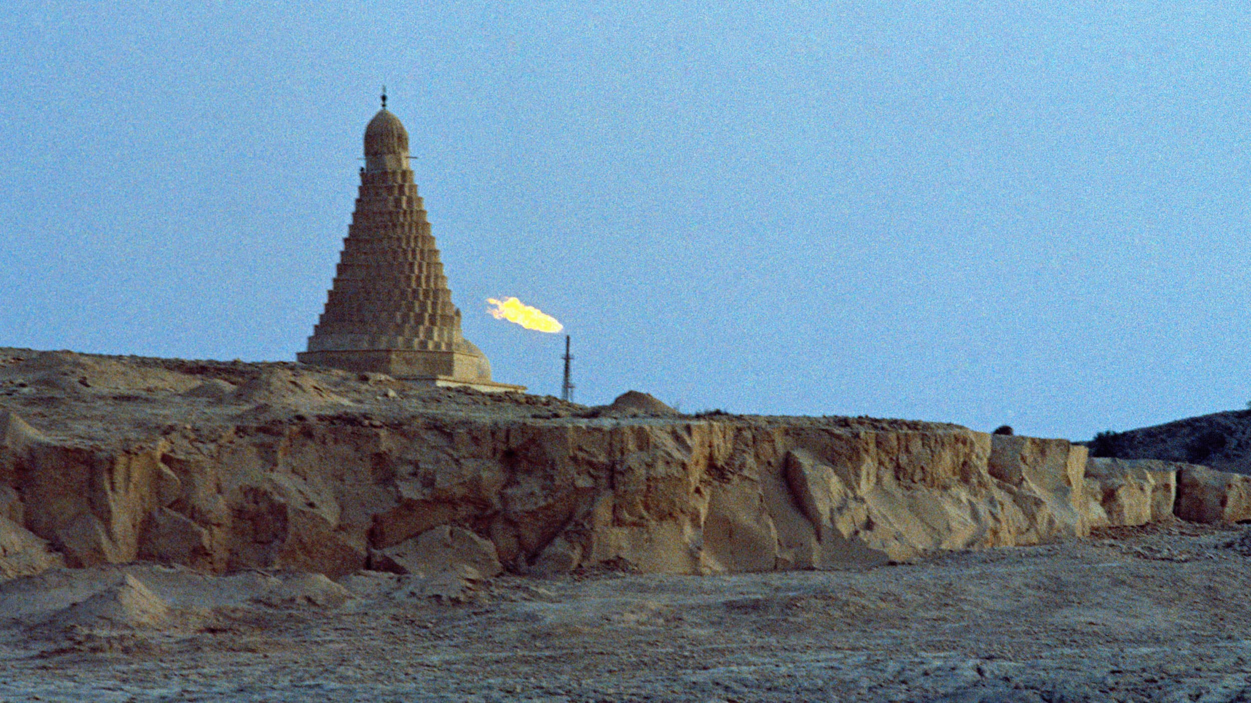 A distinctive, tiered cone-shaped shrine stands on a rocky hilltop against a blue sky, with a fiery gas flare visible nearby.