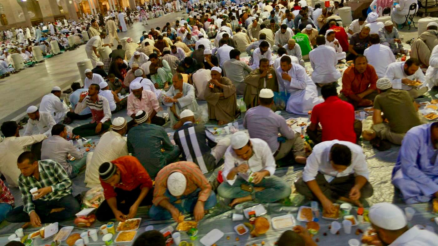 A large gathering of people seated on the ground, sharing a communal meal during a religious event, with many wearing traditional white clothing.