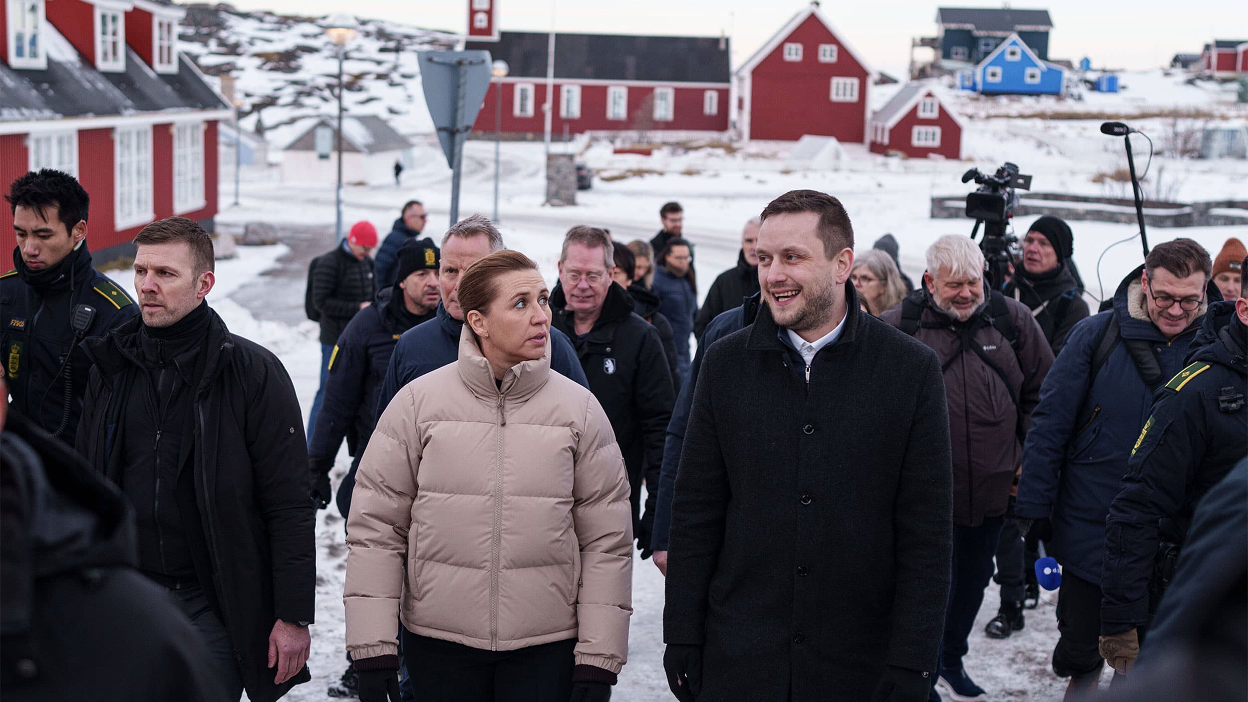 A group of people walking through a snowy village with red houses, featuring a woman in a beige puffer jacket and a man in a black coat talking in the foreground.