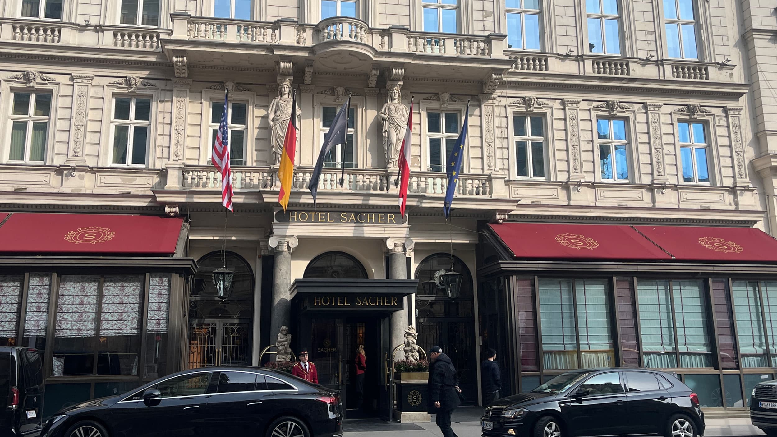 Facade of Hotel Sacher with ornate architecture, multiple flags including American and European, and a few parked cars outside.
