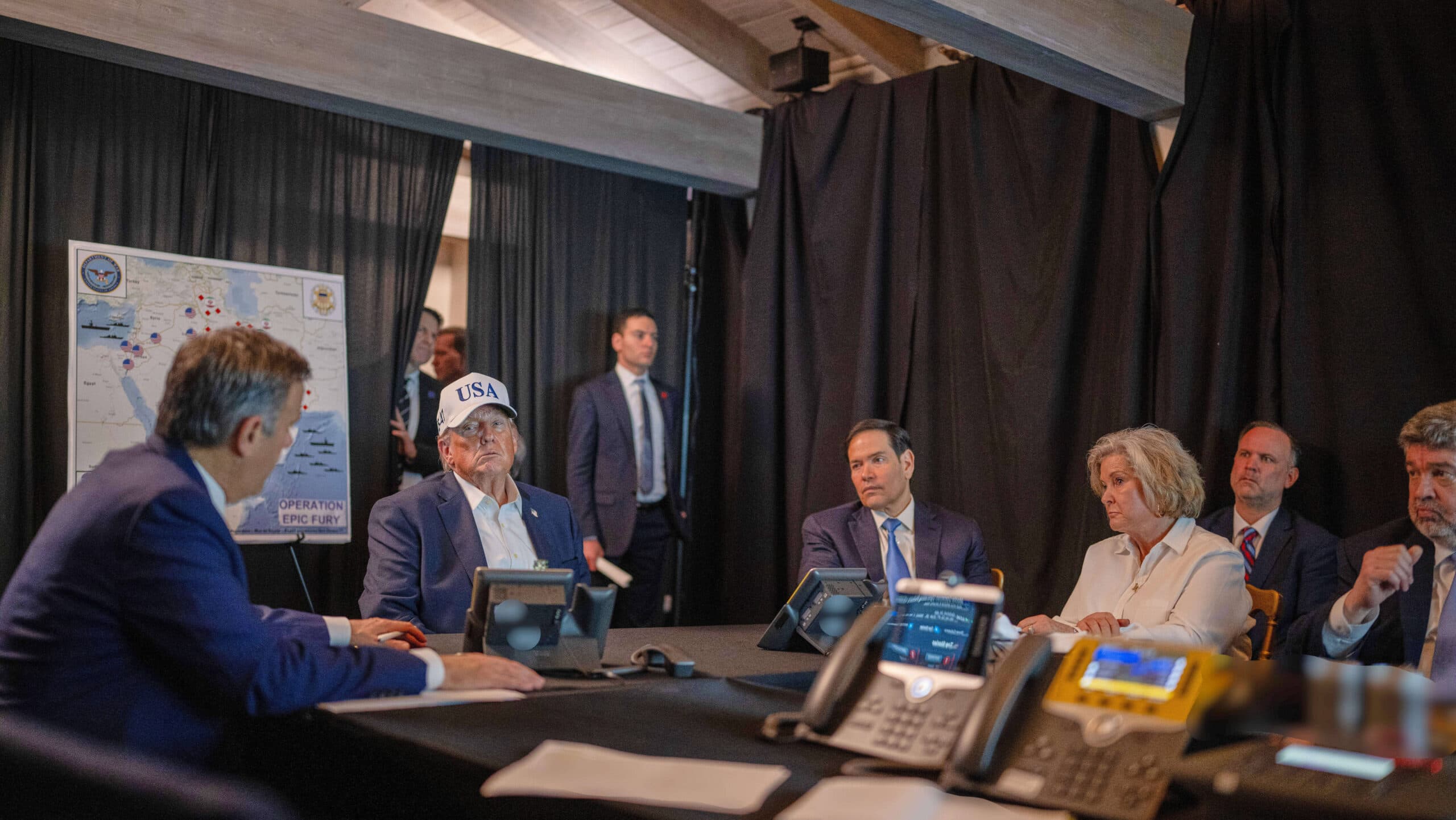 Several individuals in suits sitting around a conference table with phones, one wearing a USA cap, engaged in discussion with a large map labeled "Operation Epic Fury" in the background.