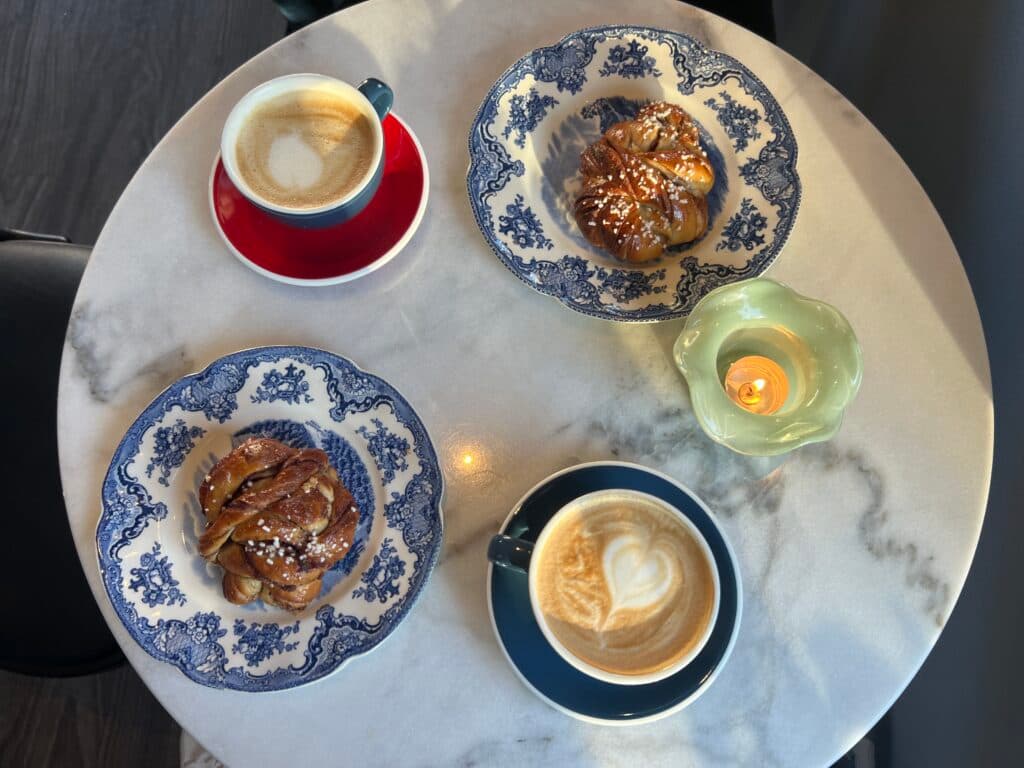 Two cups of coffee with latte art and two pastries on patterned plates beside a lit candle on a marble table.