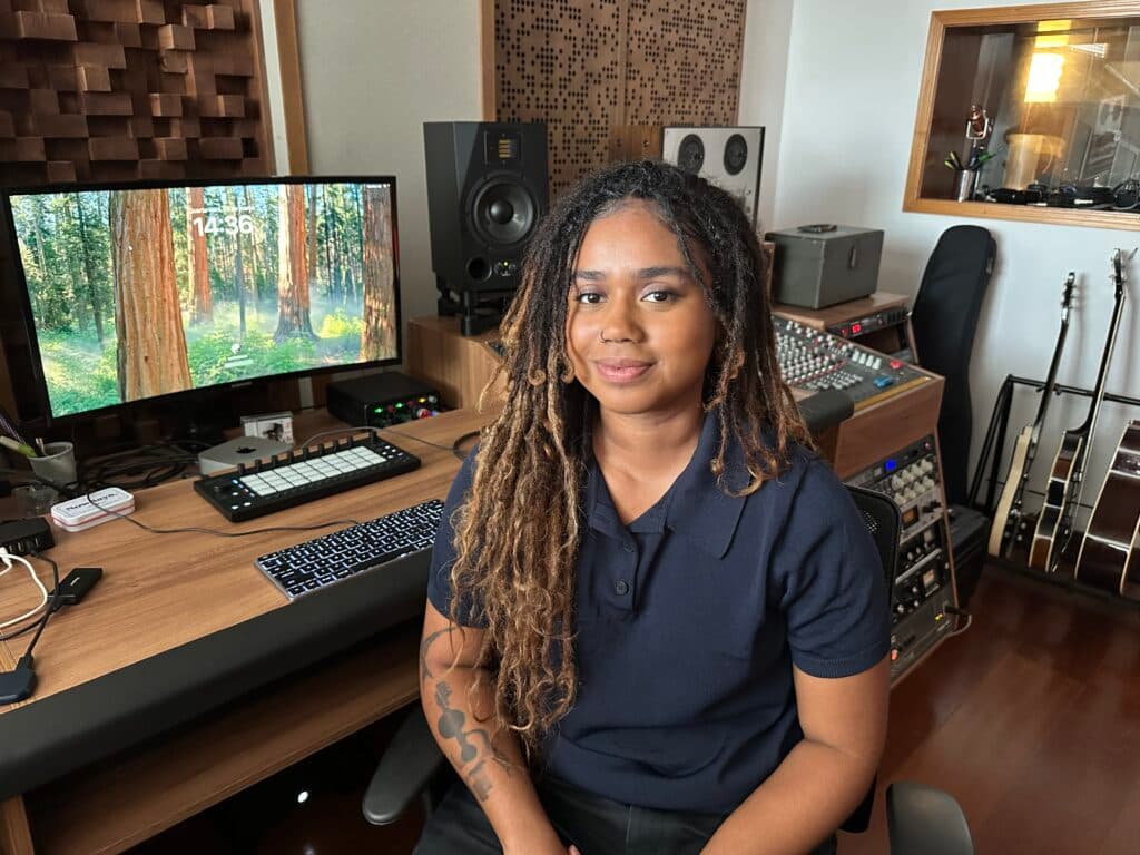A person with long locs is seated in a music studio surrounded by audio equipment and guitars.
