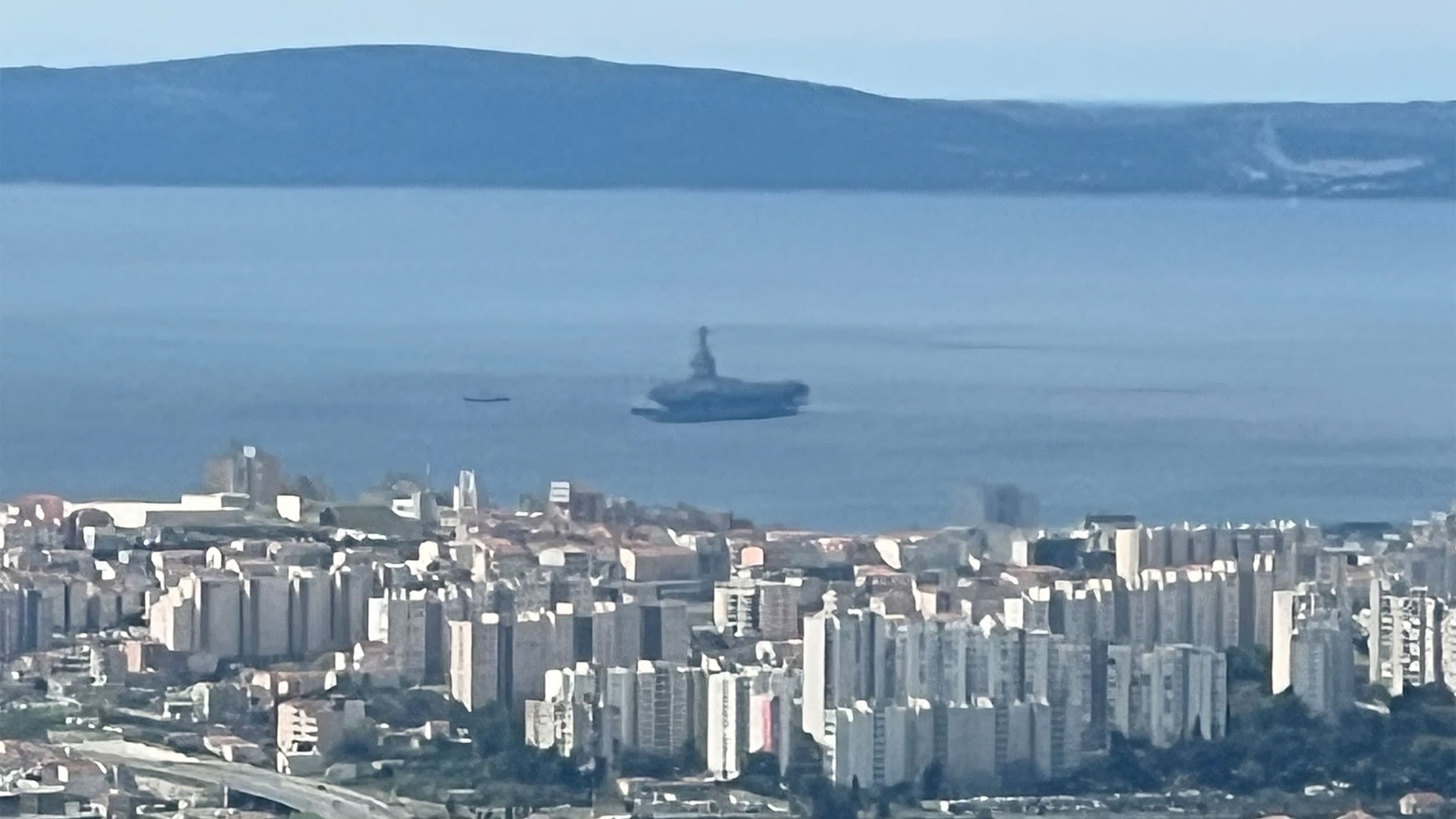 An aircraft carrier appears to be floating above the water in an optical illusion caused by atmospheric conditions, seen from a distance with a cityscape in the foreground and a hilly horizon in the background.