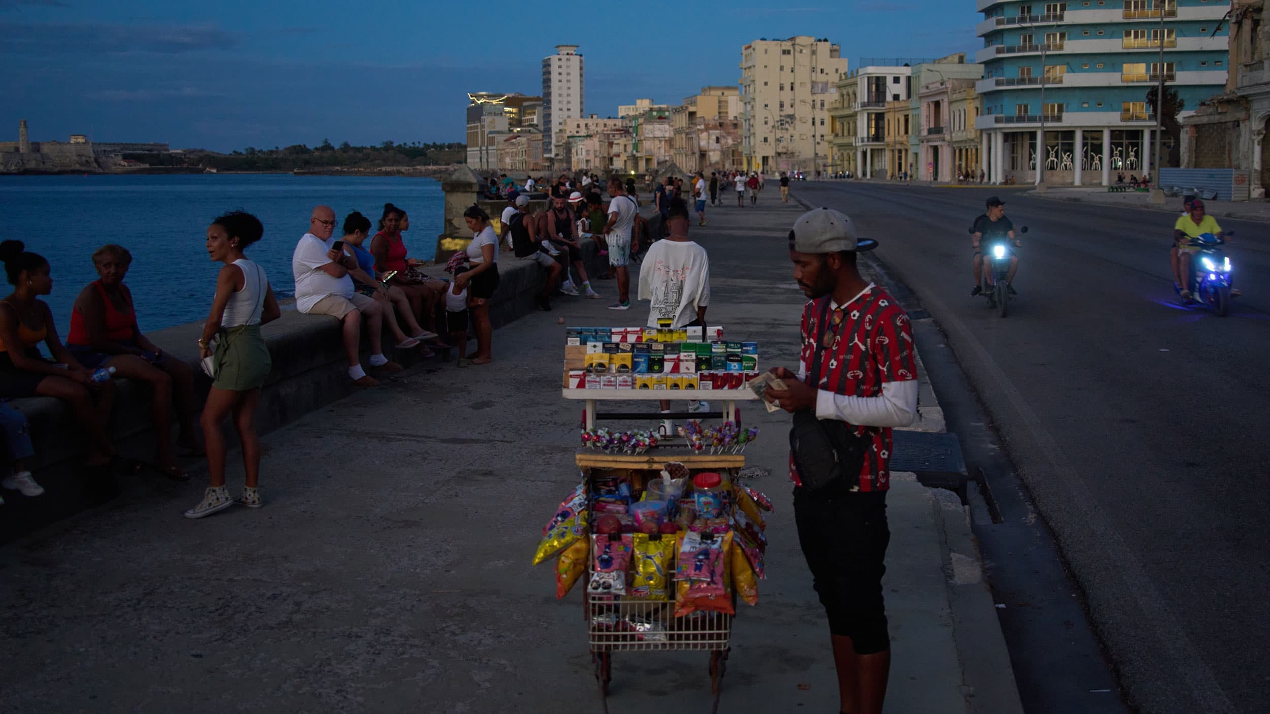A street vendor with a cart full of assorted goods, including snacks and cigarettes, stands on a waterfront promenade as people sit along a low wall by the sea. The street is lined with people and buildings in the background, while two motorbikes with headlights on pass by on the road.
