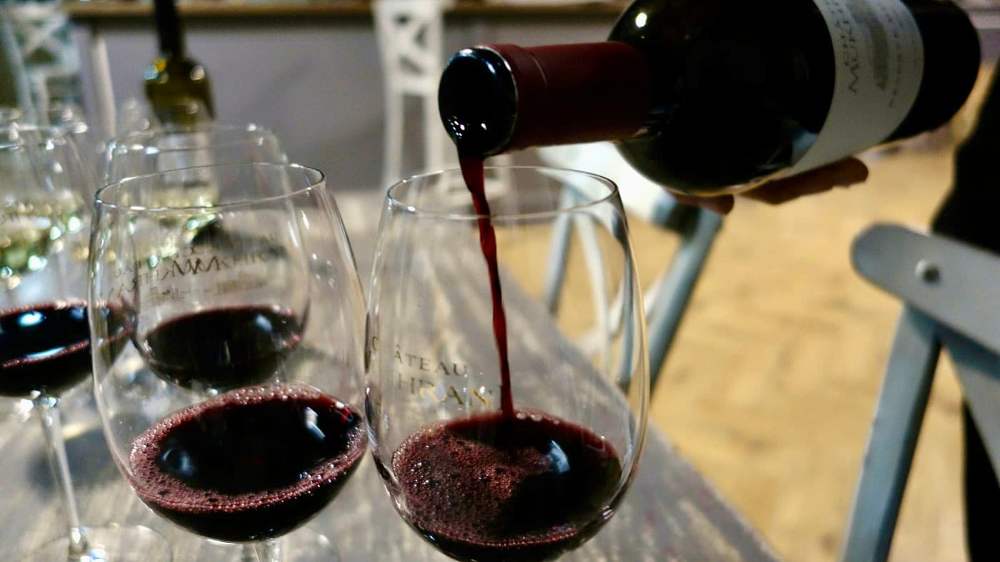 A person pouring red wine into a glass, with other filled glasses in the background, on a wooden table.