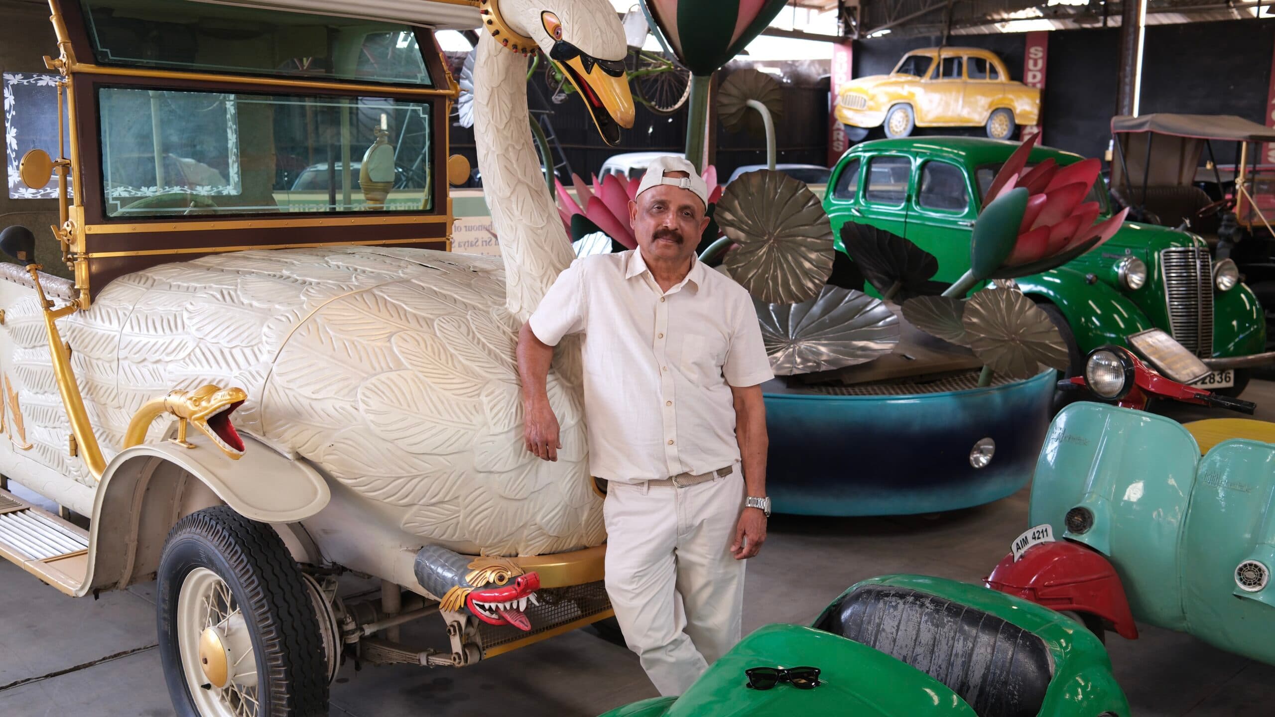 A man wearing a white outfit is standing next to a vintage car designed to resemble a swan, with ornate decorations and a snake ornament on the fender. The background features other brightly colored, uniquely designed vehicles.
