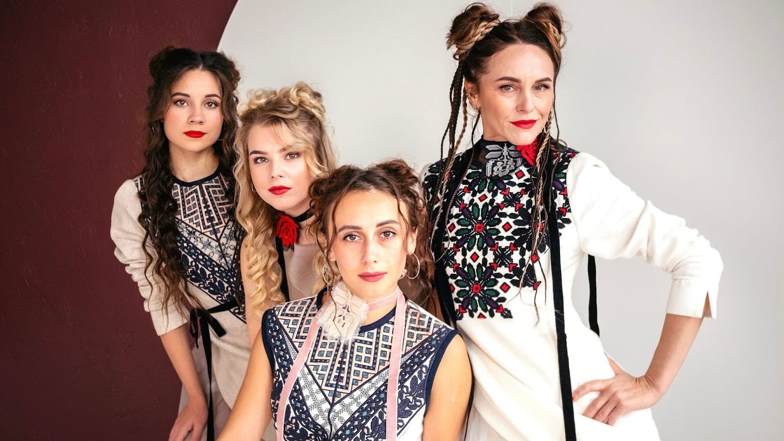 Four women in embroidered clothing pose against a light background, each with unique hairstyles and wearing red lipstick.