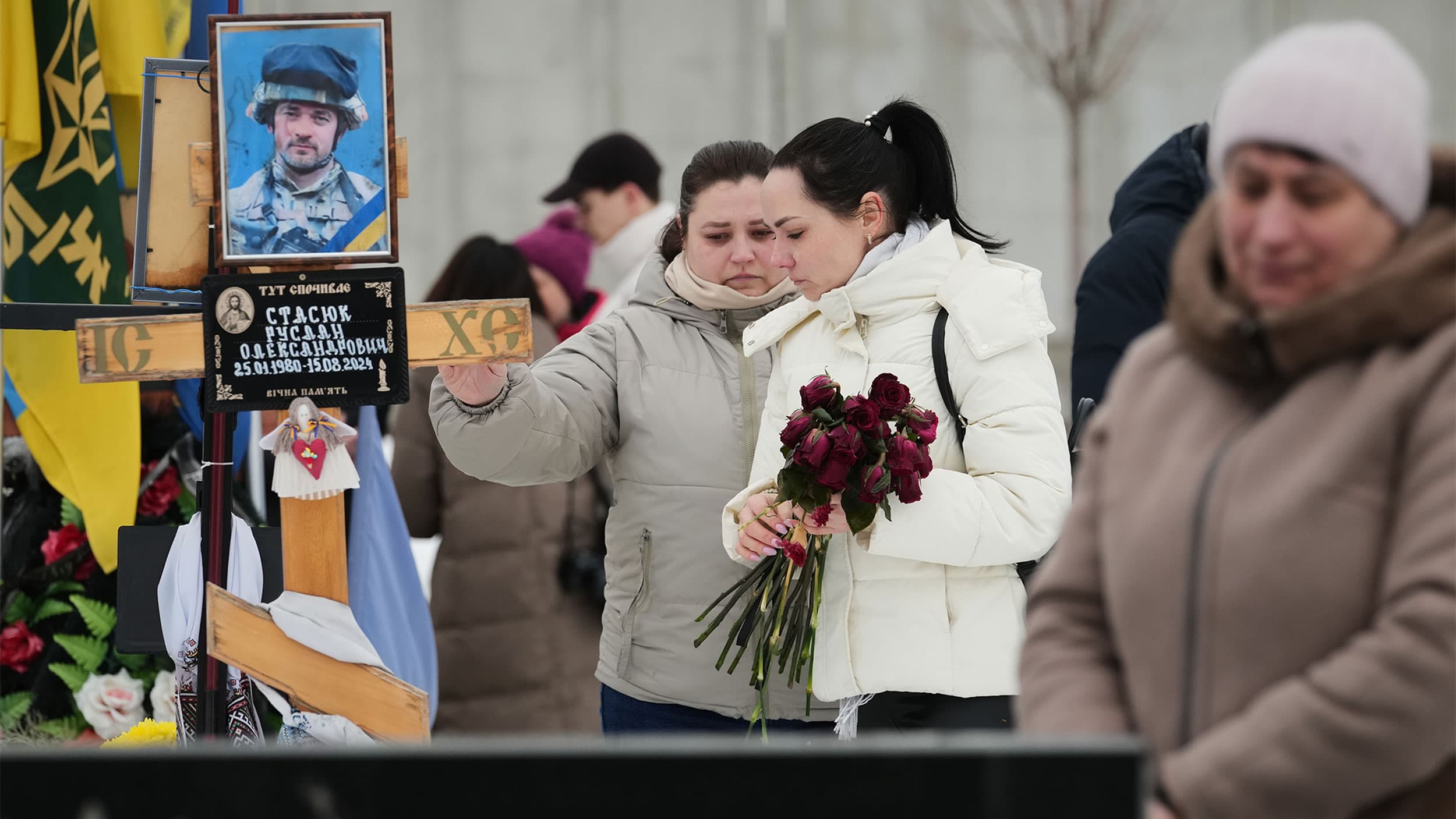Two women in winter clothing stand at a grave holding flowers, with a memorial portrait in the background.