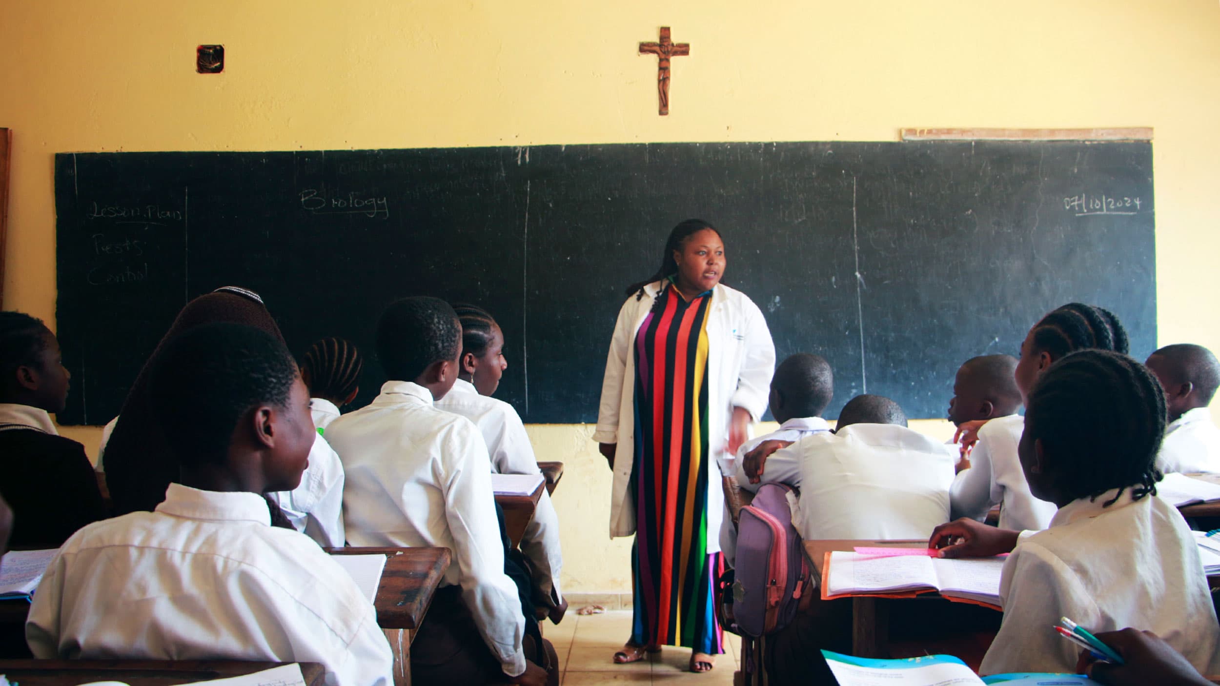A teacher in a colorful dress stands in front of a classroom, with students sitting at desks and a blackboard behind her.