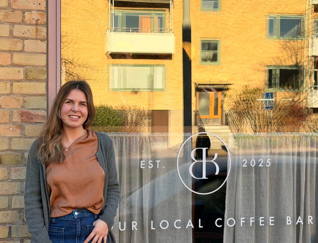 A smiling woman stands outside a brick building beside a window displaying a coffee bar logo and the text "OUR LOCAL COFFEE BAR EST. 2025."