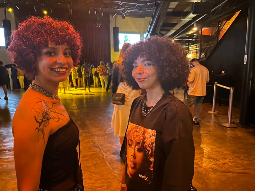 Two people with curly hair smiling in a dimly lit venue, with others in the background on a dance floor.