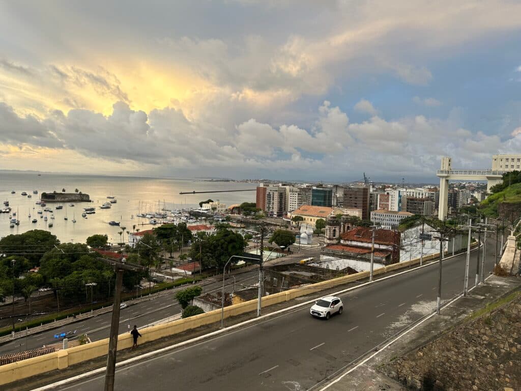 Coastal city view at sunset with a road, buildings, harbor, and scattered boats under a partly cloudy sky.