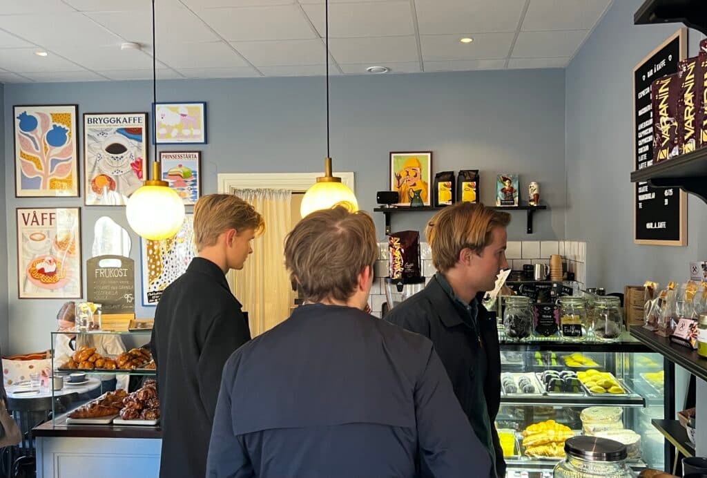 Three people stand inside a cafe, looking at a display case with pastries and desserts.