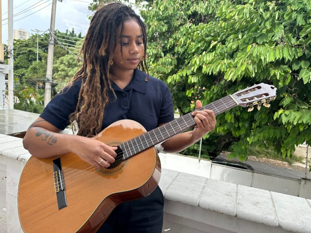 A person with long braided hair plays an acoustic guitar outdoors near green foliage.