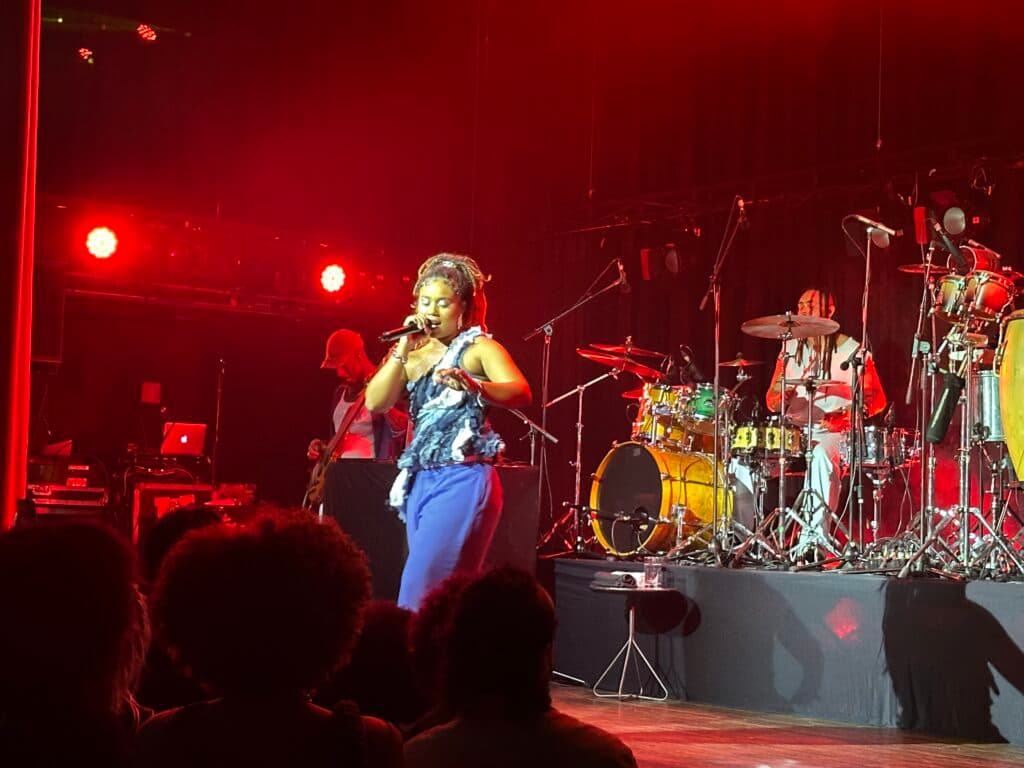 A singer on stage performs under red lights with a band in the background.