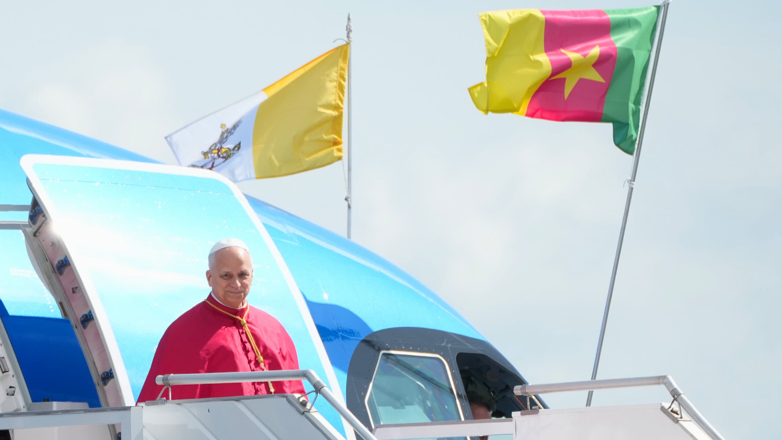 A man in religious attire stands at the top of airplane stairs, with Vatican and Cameroon flags visible above.